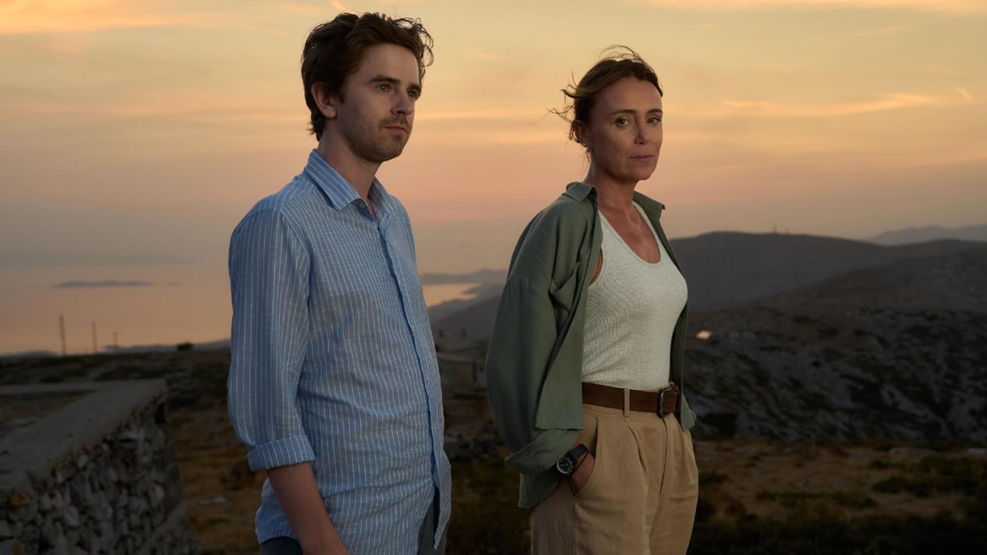 Ator Freddie Highmore numa camisa azul e atriz Keeley Hawes numa camisola branca contra um cenário montanhoso