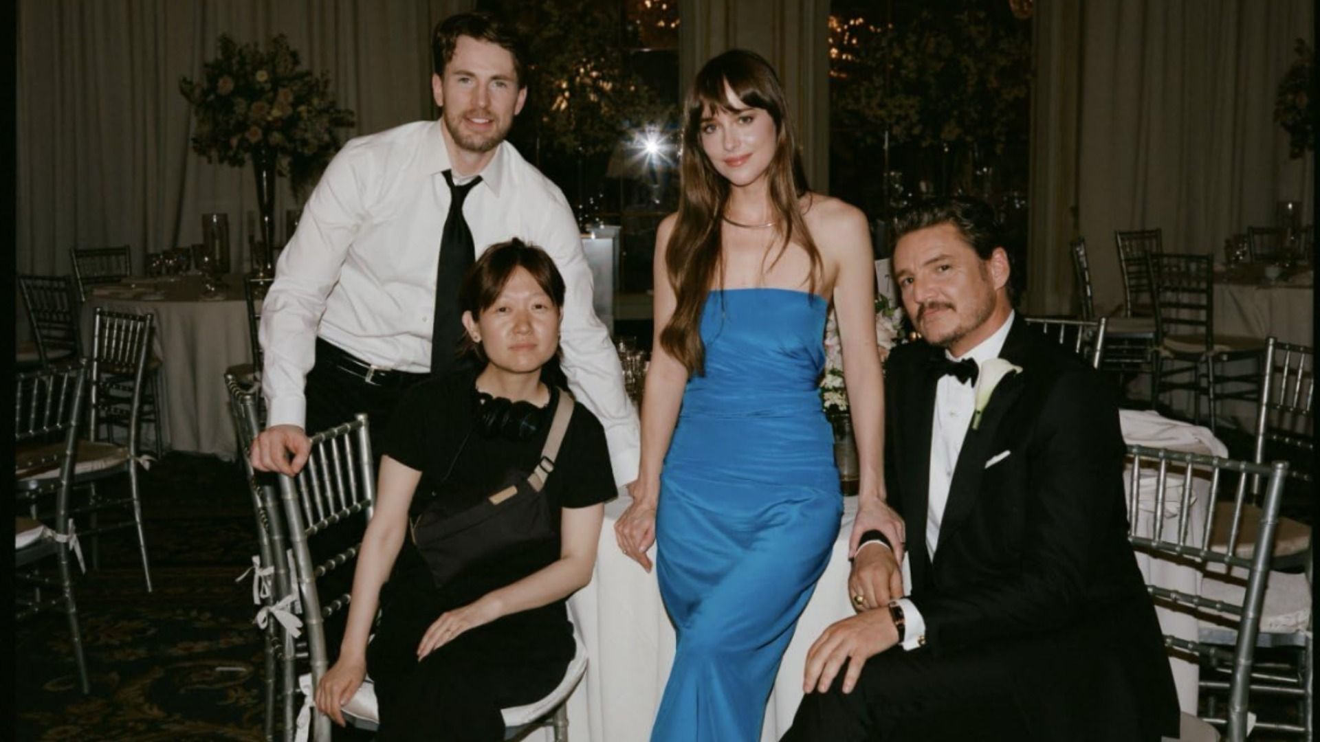 Atores Dakota Johnson, Chris Evans e Pedro Pascal sentados em traje de gala numa mesa branca