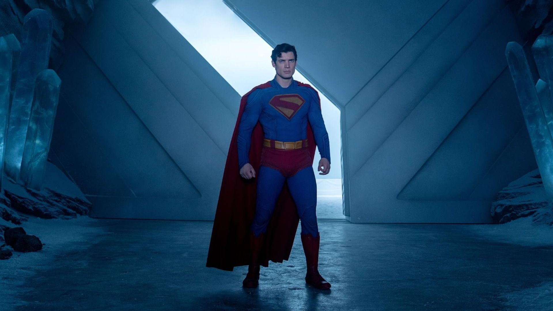 O ator David Corenswet num fato de super-herói azul e vermelho está junto a grandes portões
