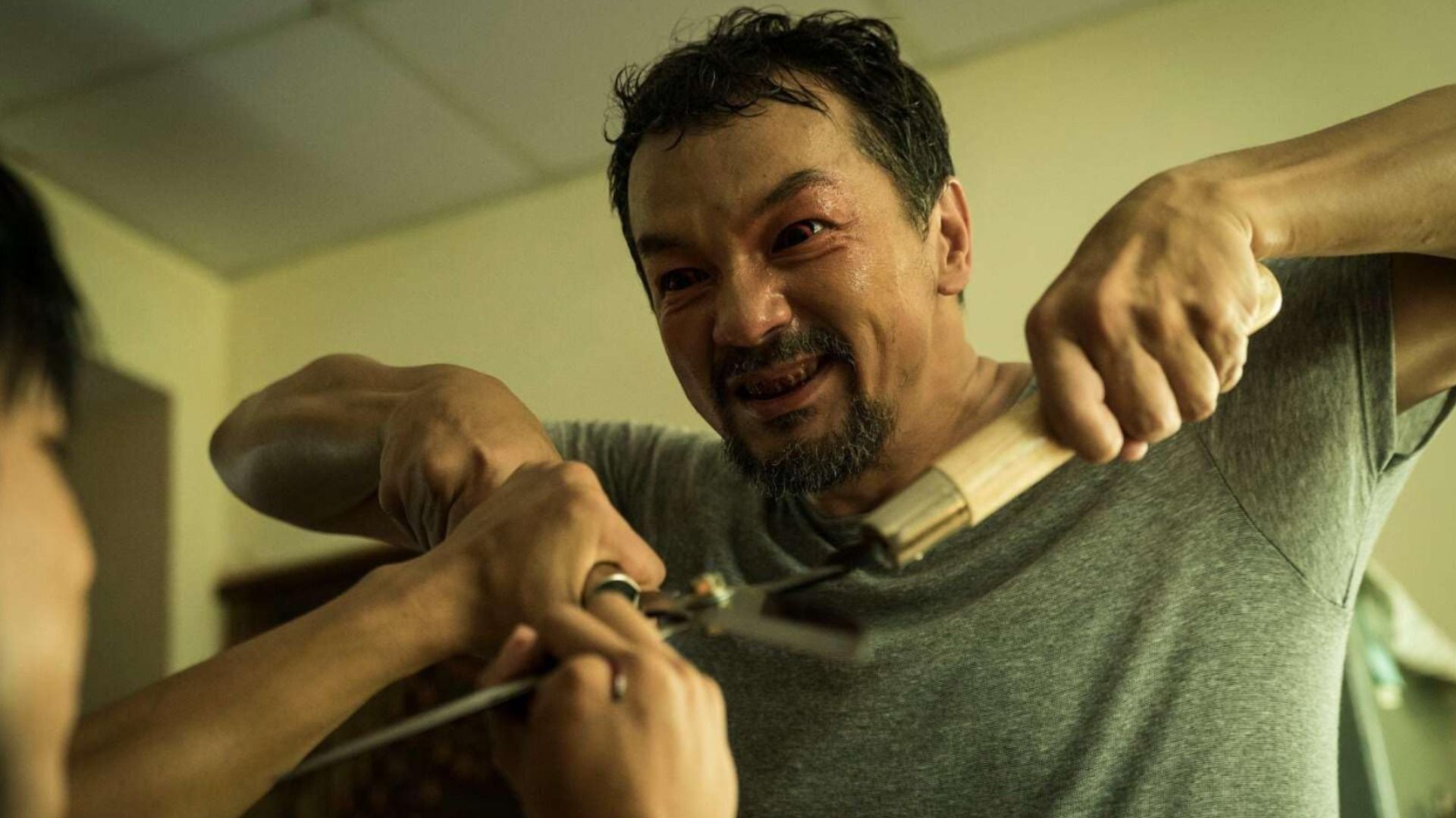 Um homem numa t-shirt cinzenta com olhos vermelhos segura uma tesoura de poda