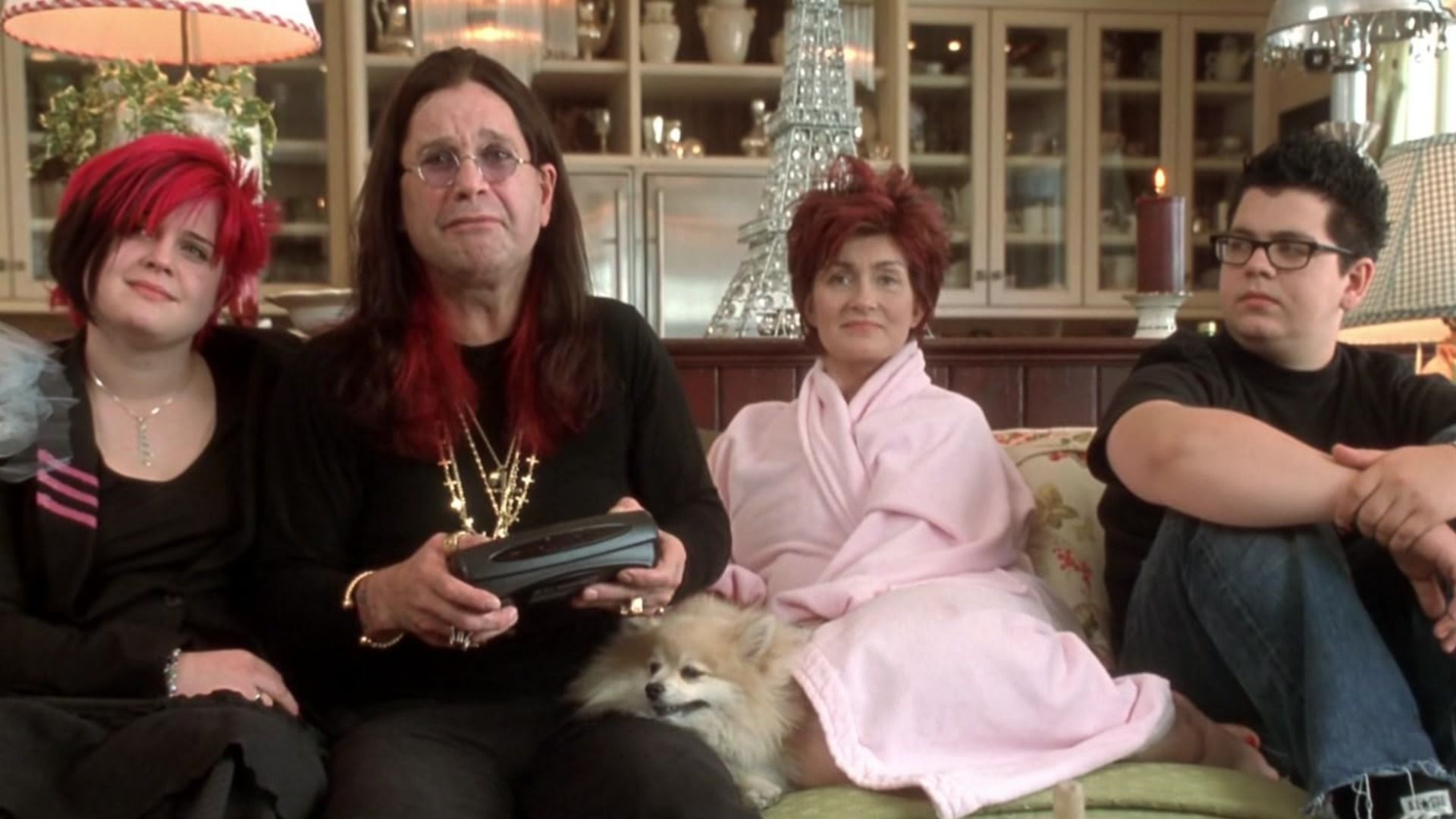 O cantor Ozzy Osbourne num fato preto com correntes sentado num sofá com a mulher e filhos