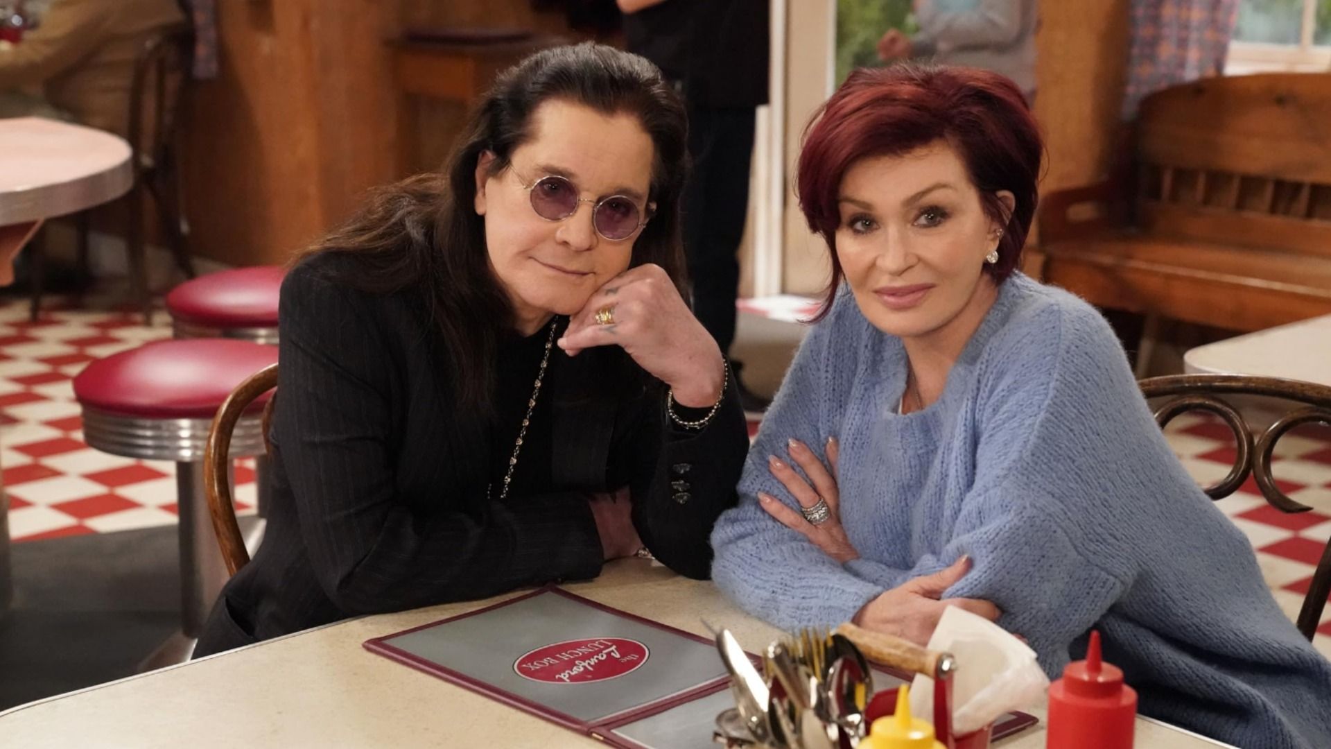 O cantor Ozzy Osbourne de óculos e casaco preto sentado à mesa com a mulher