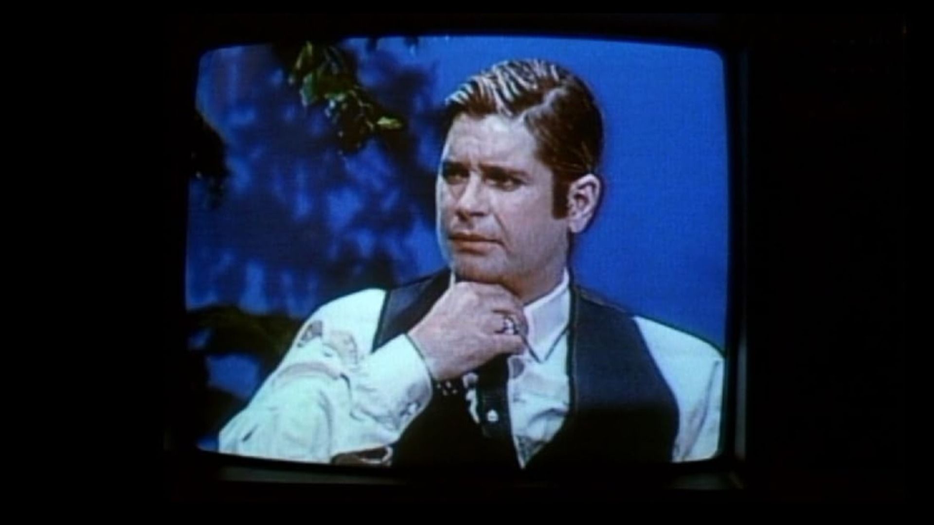 O cantor Ozzy Osbourne de cabelo curto sentado à mesa vestindo camisa branca e colete preto