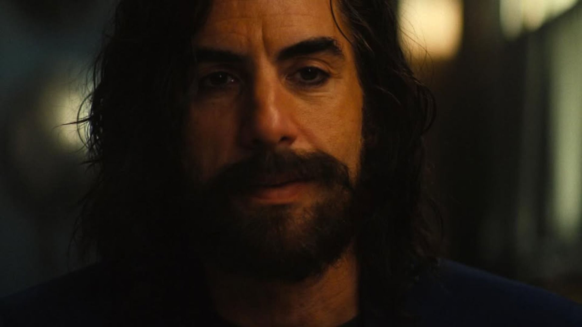 O ator Sacha Baron Cohen com barba e cabelo comprido está de pé contra um fundo desfocado