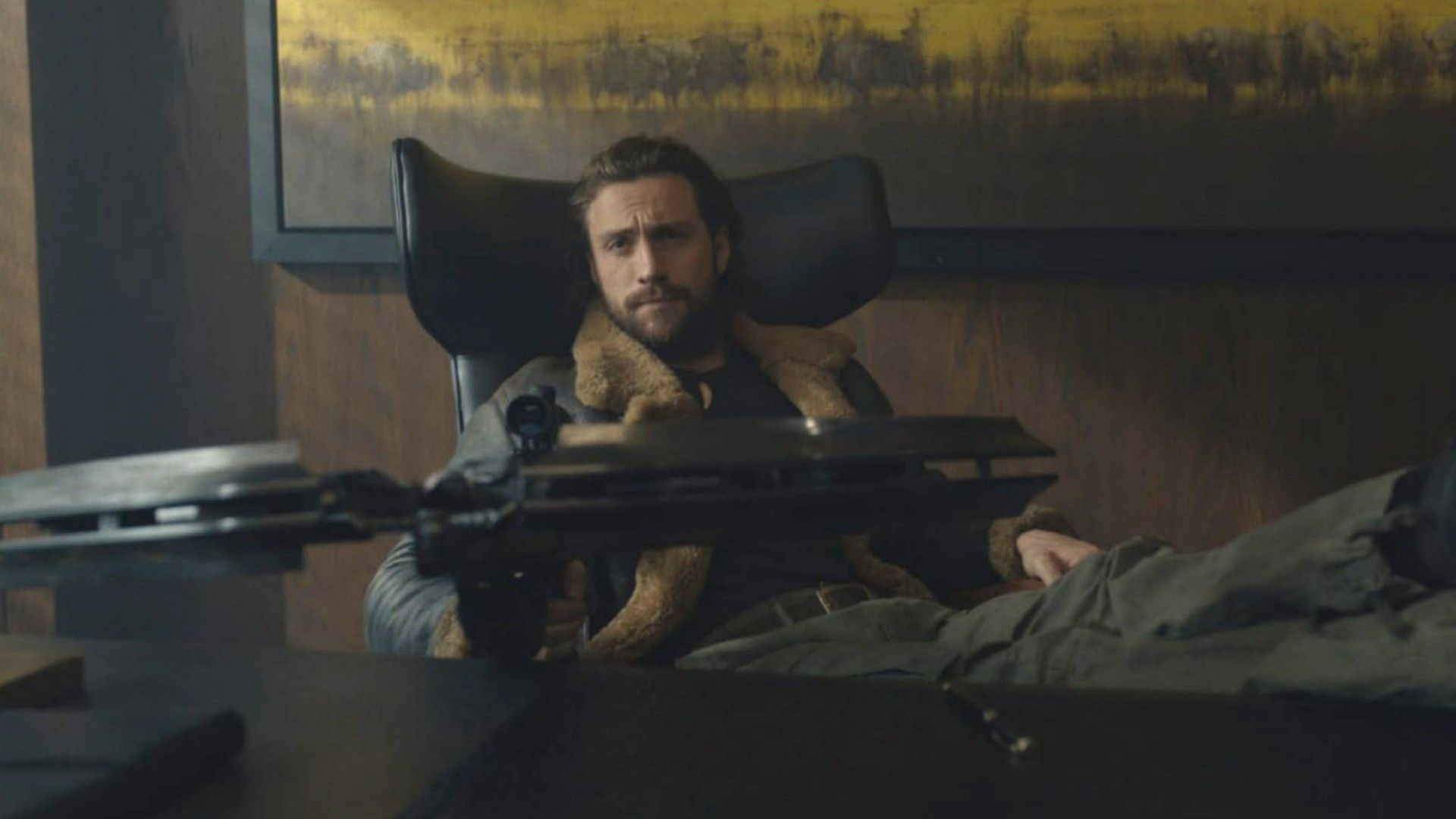 O ator Aaron Taylor-Johnson está sentado a uma mesa com uma besta nas mãos