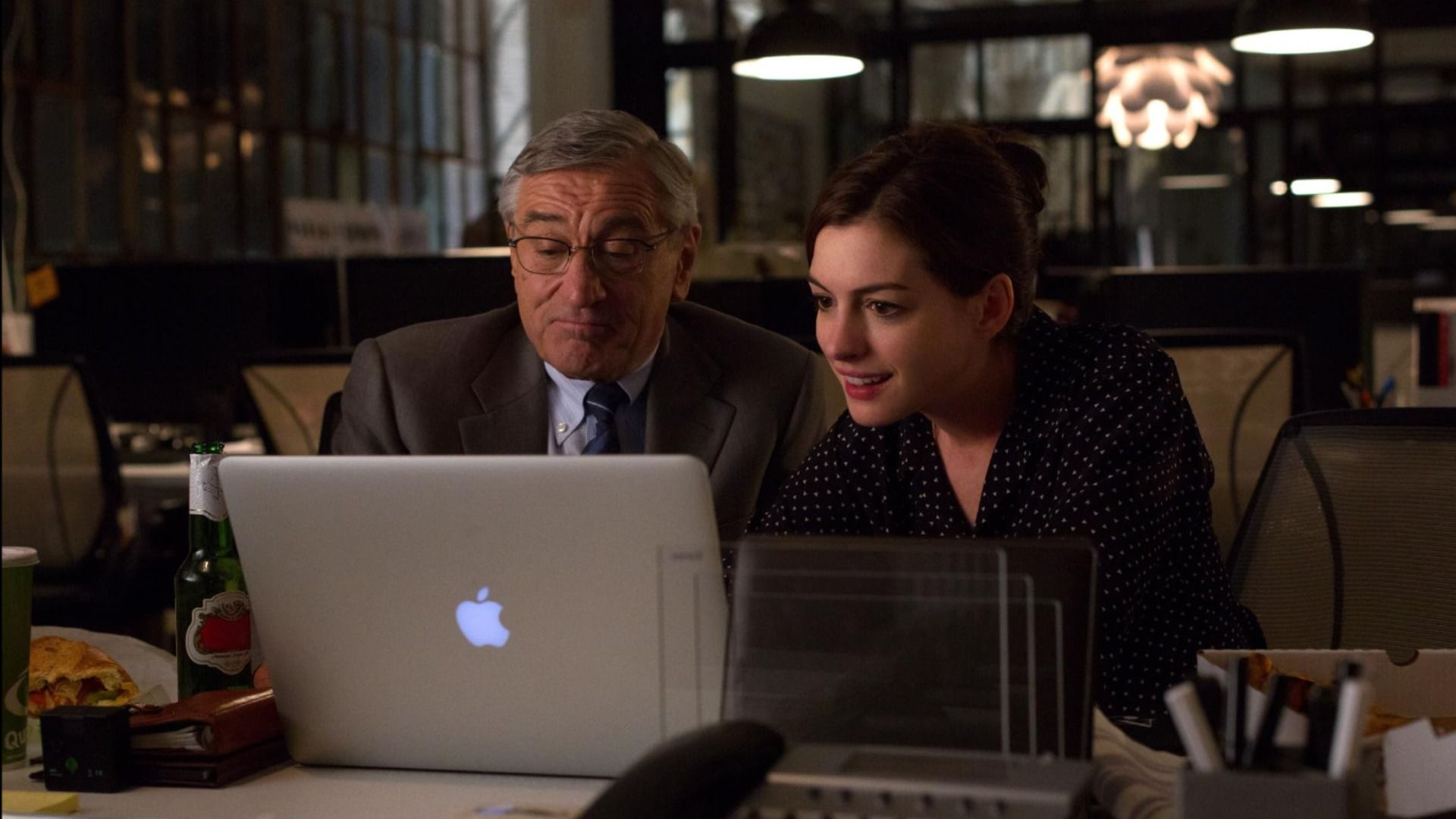 Ator Robert De Niro num casaco de fato cinzento e a atriz Anne Hathaway num vestido preto sentados à mesa