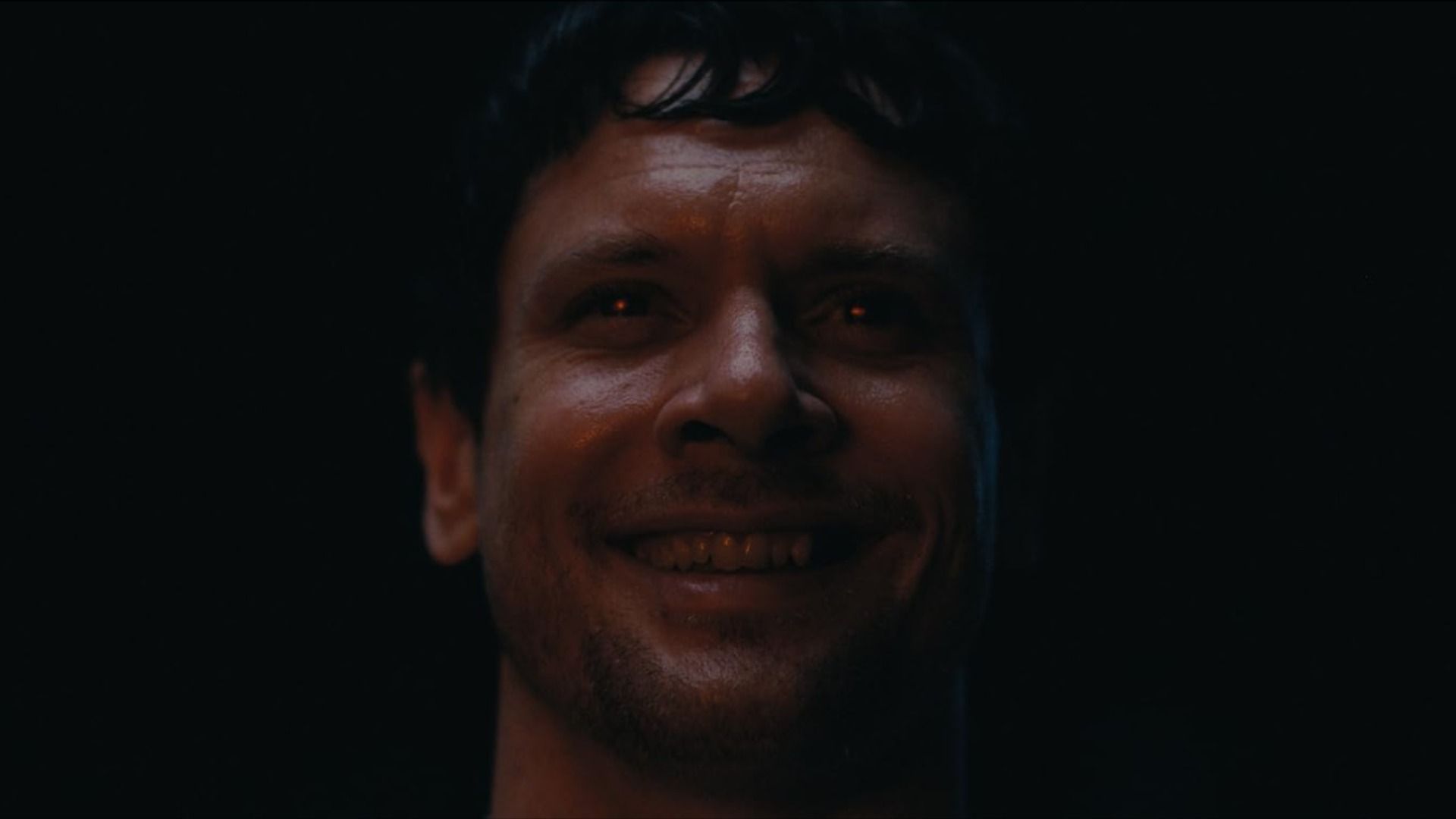 Um homem sorri contra um fundo preto com um brilho vermelho nos olhos