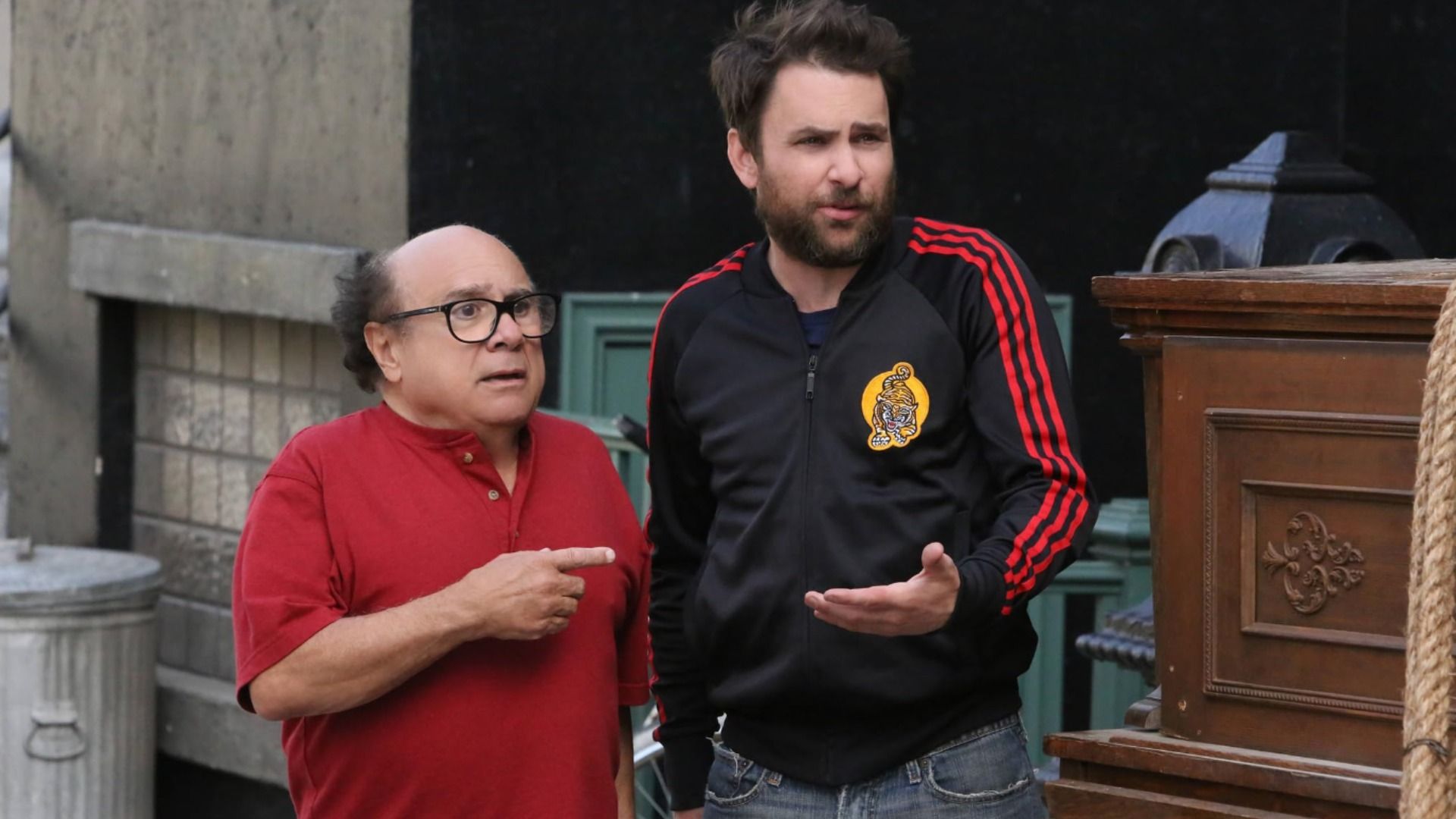 O ator Danny DeVito numa camisa vermelha aponta o dedo para um homem com roupa desportiva
