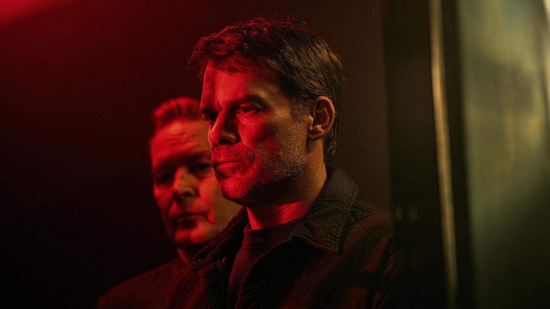 O ator Michael C. Hall está de perfil numa iluminação vermelha com parte do rosto de um homem visível atrás dele