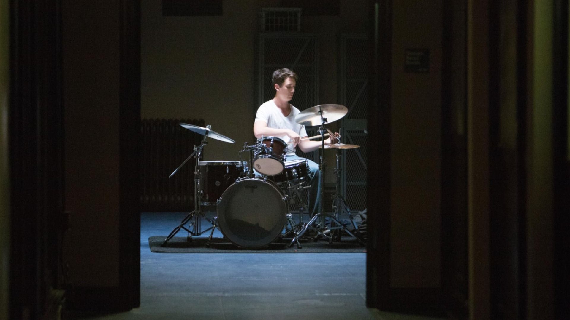 Iluminação suave, o ator Miles Teller toca bateria numa t-shirt branca