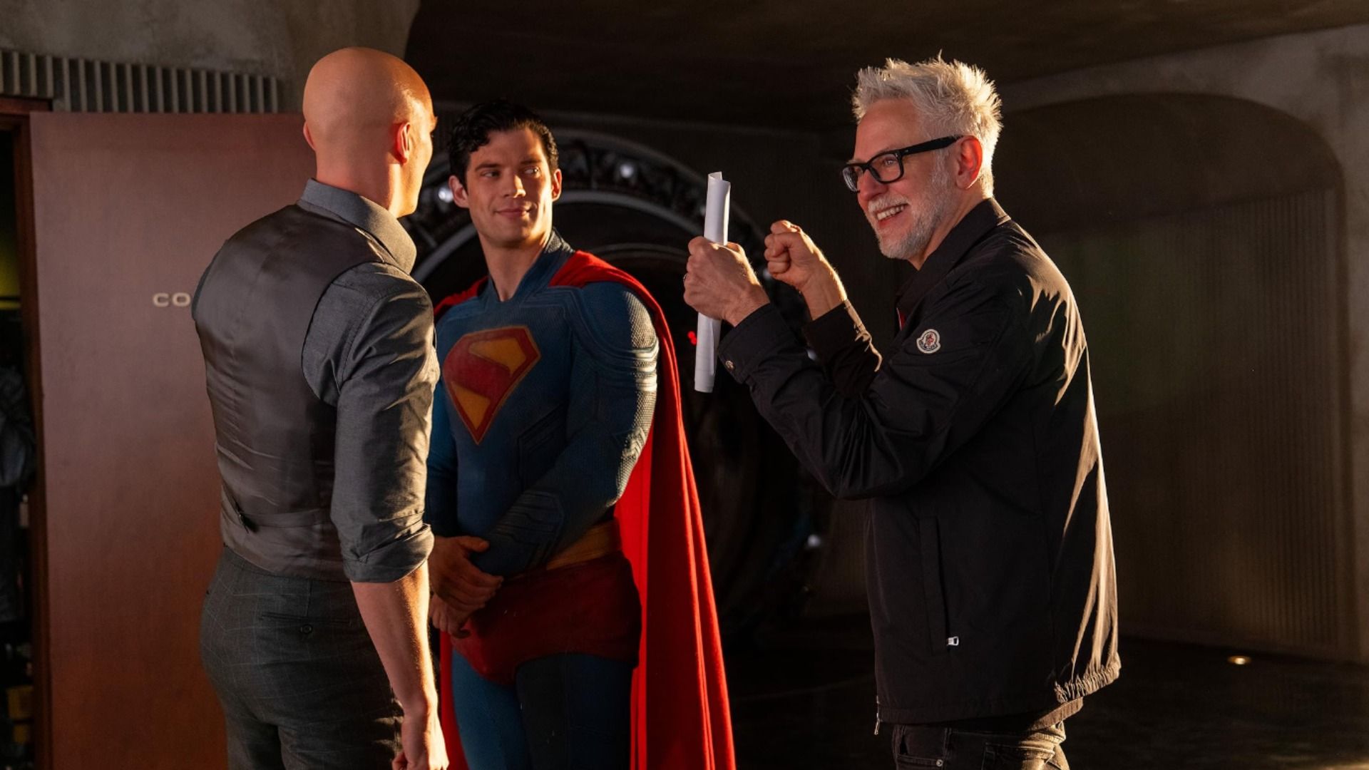 O realizador James Gunn segurando papéis conversa com o ator David Corenswet no fato do Superman e com Nicholas Hoult que está careca