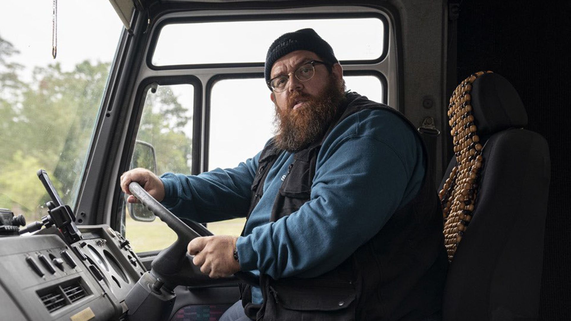 Nick Frost usando óculos e um chapéu está sentado ao volante de um camião, olhando para a câmara