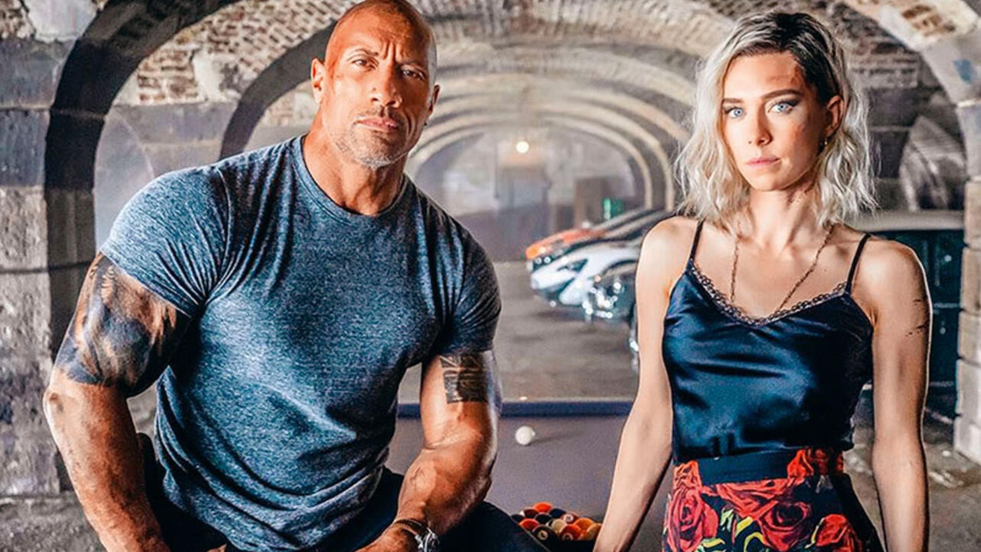Dwayne Johnson numa t-shirt justa e Vanessa Kirby numa camisola de alças e saia estampada posam contra um cenário de arcos e carros desportivos