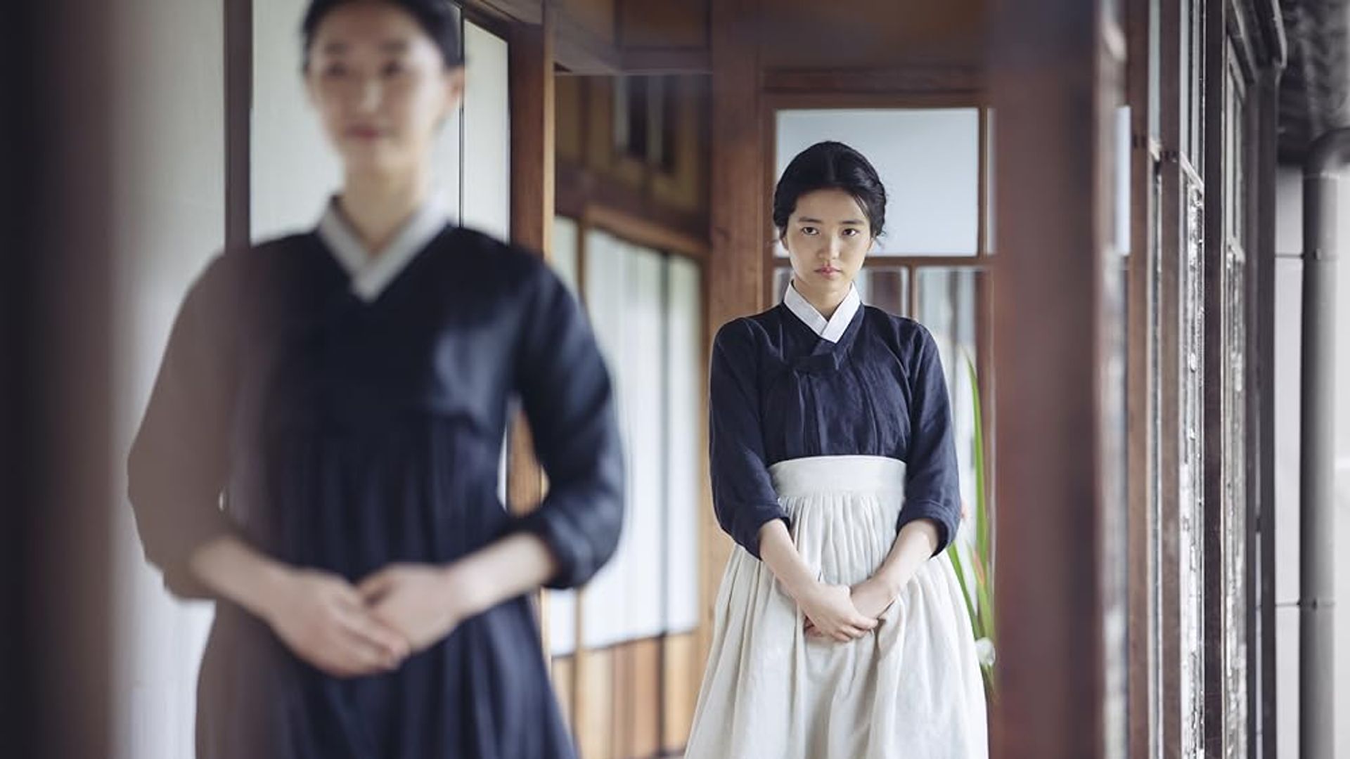 Uma rapariga num hanbok preto com avental branco está no corredor, olhando em frente com expressão séria