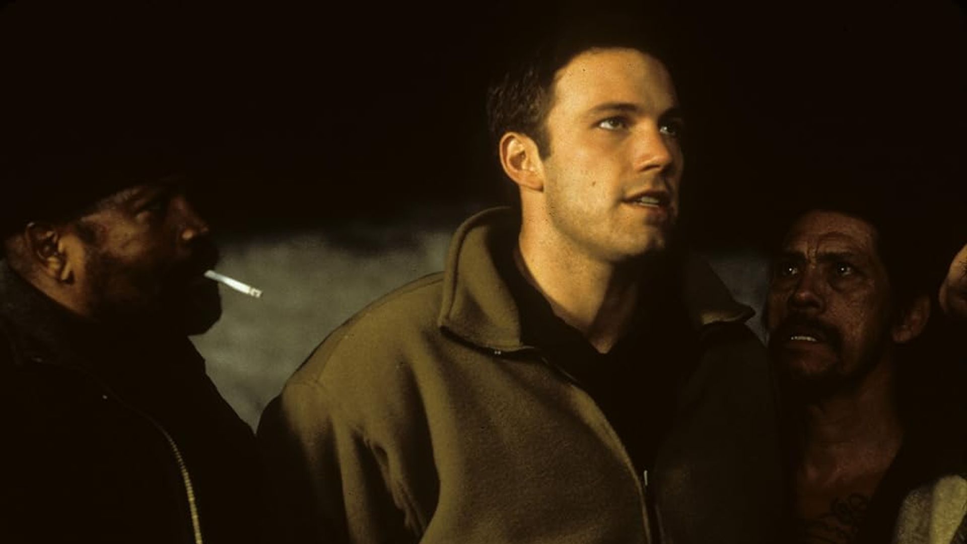 Ben Affleck com um casaco bege entre duas pessoas, uma das quais está a fumar, num ambiente mal iluminado