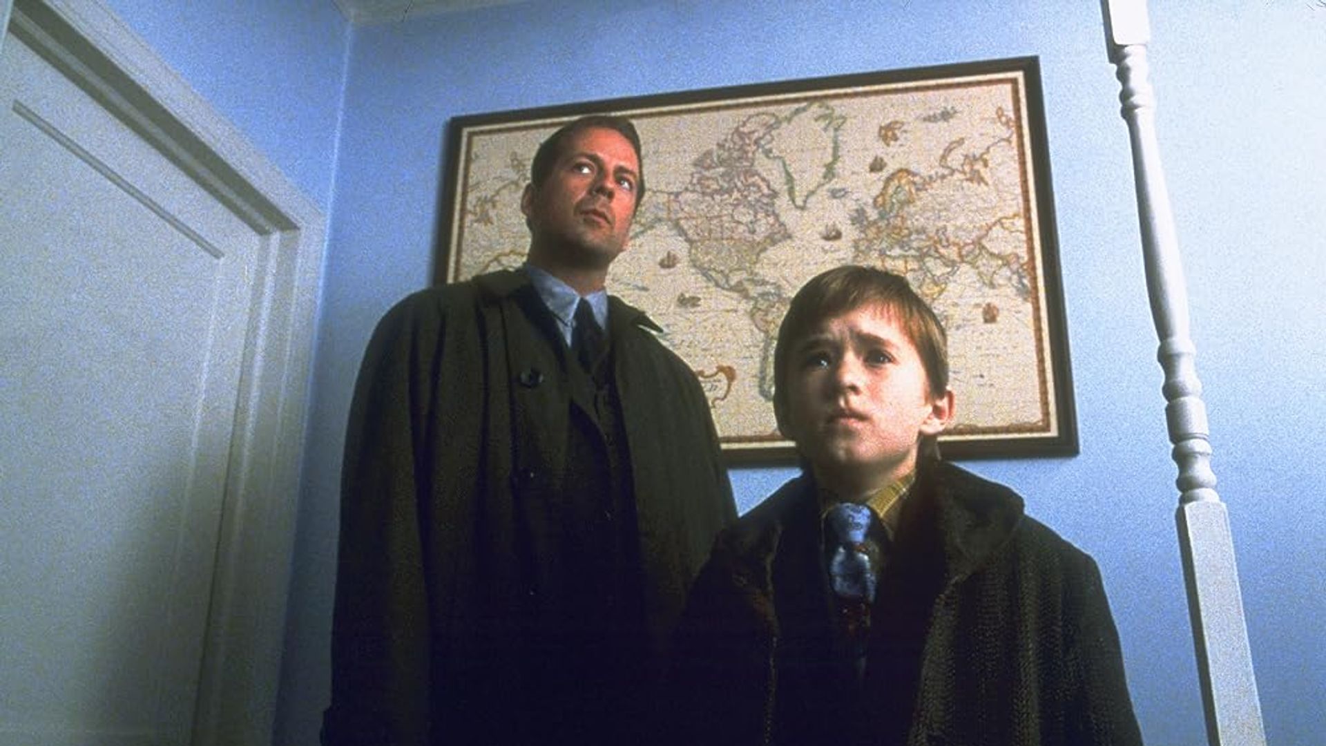 Bruce Willis e Haley Joel Osment num corredor com um mapa-mundo pendurado na parede atrás deles
