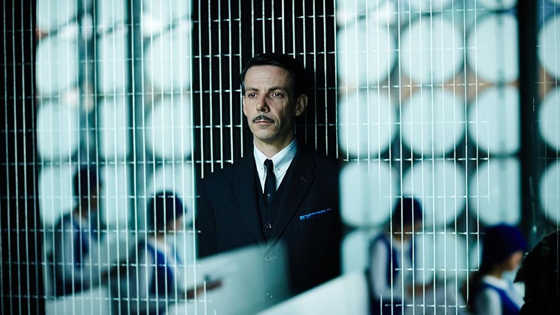 Ethan Hawke de fato e bigode atrás de uma divisória de rede com pessoas uniformizadas a trabalhar em secretárias ao fundo