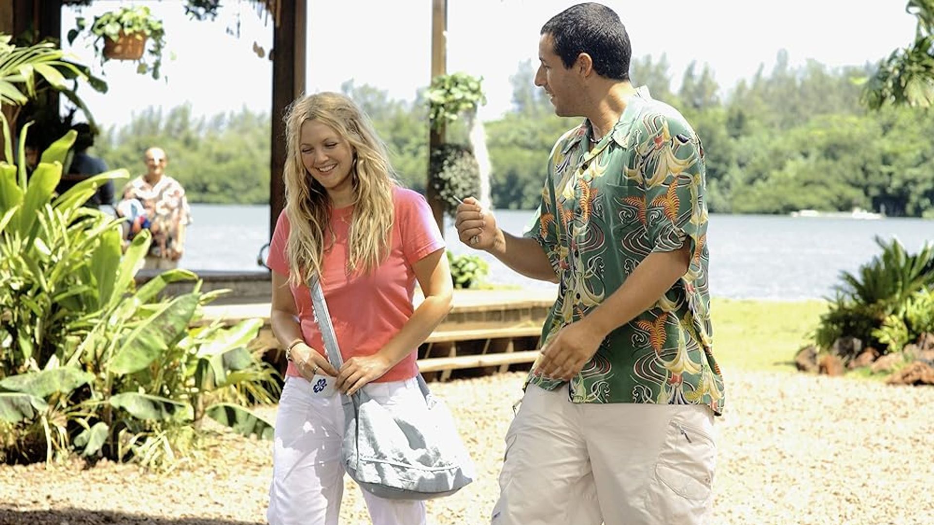 Drew Barrymore e Adam Sandler a conversar numa praia ensolarada com vegetação e água ao fundo