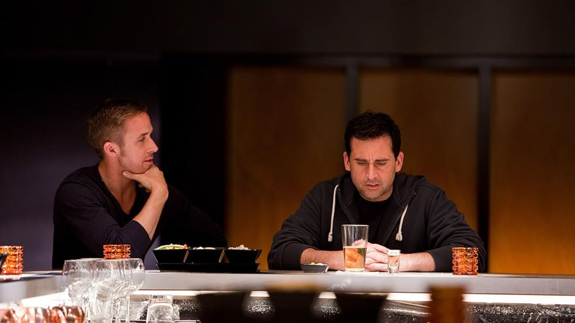 Ryan Gosling e Steve Carell sentados ao balcão de um bar, um olhando pensativamente para o copo, o outro a observar