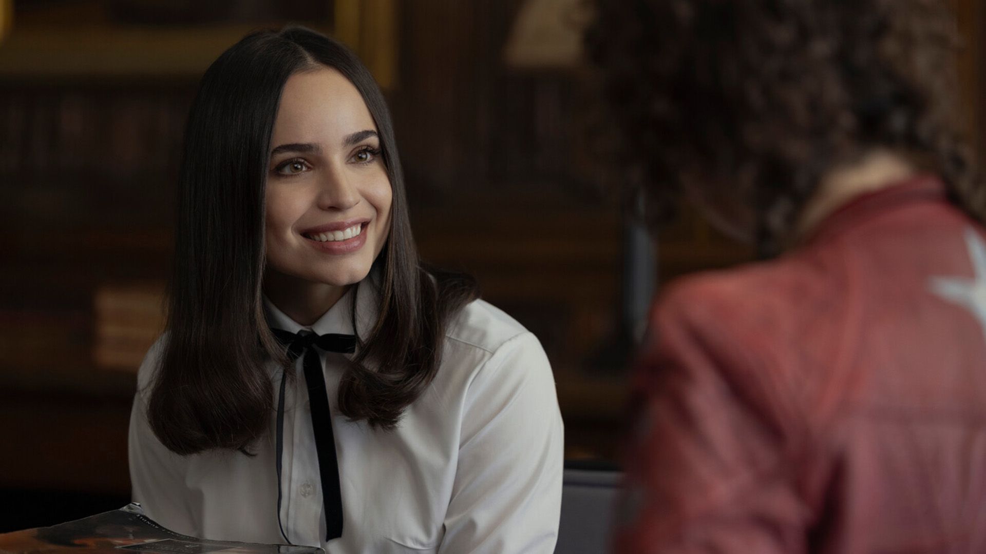 Sofia Carson numa blusa branca com um laço preto no pescoço sorrindo amplamente