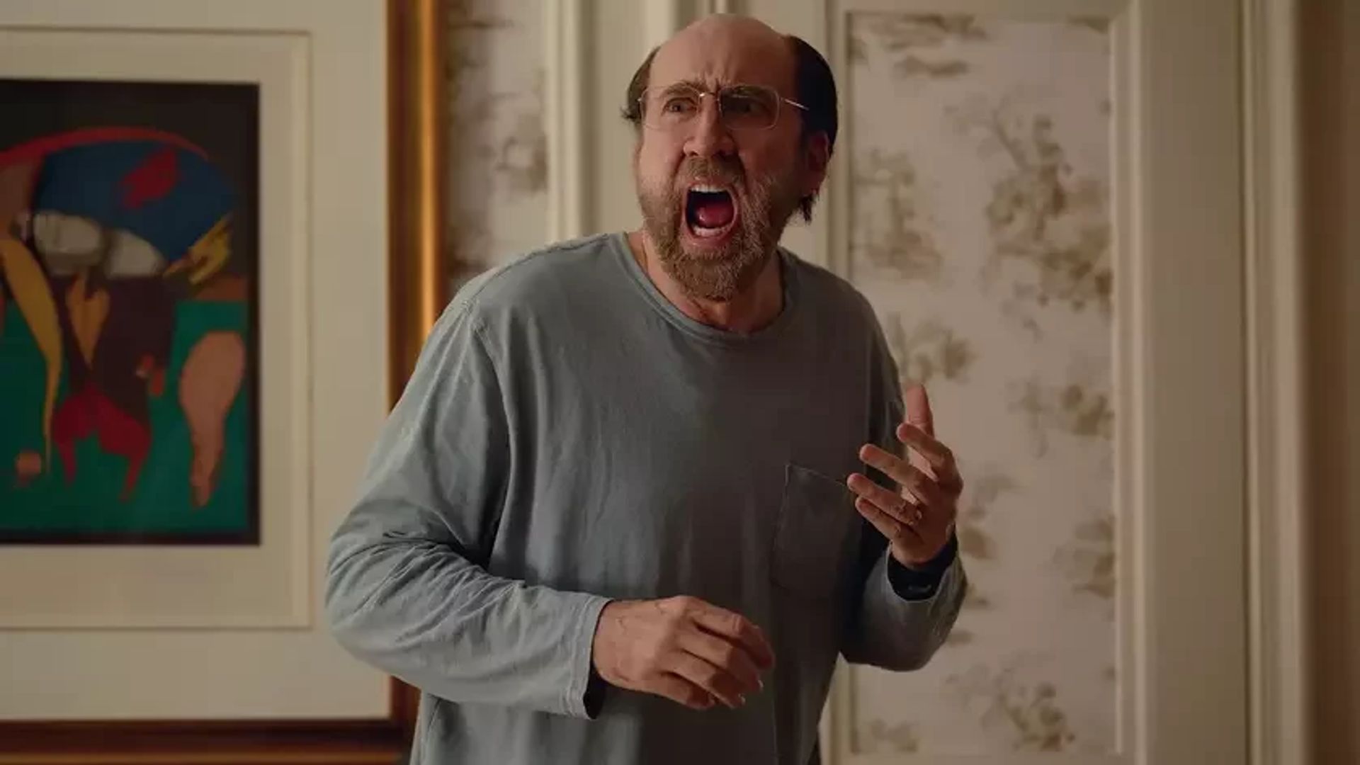 Nicolas Cage usando óculos e uma camisa cinzenta de manga comprida gritando enquanto está de pé numa sala
