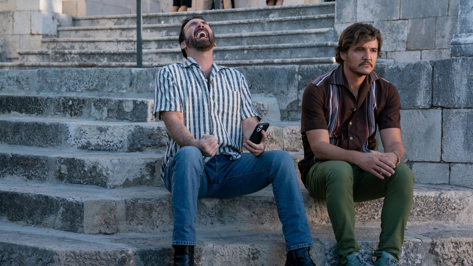 Nicolas Cage e Pedro Pascal sentados em degraus de pedra: um ri com um telefone na mão, o outro olha pensativamente para o lado