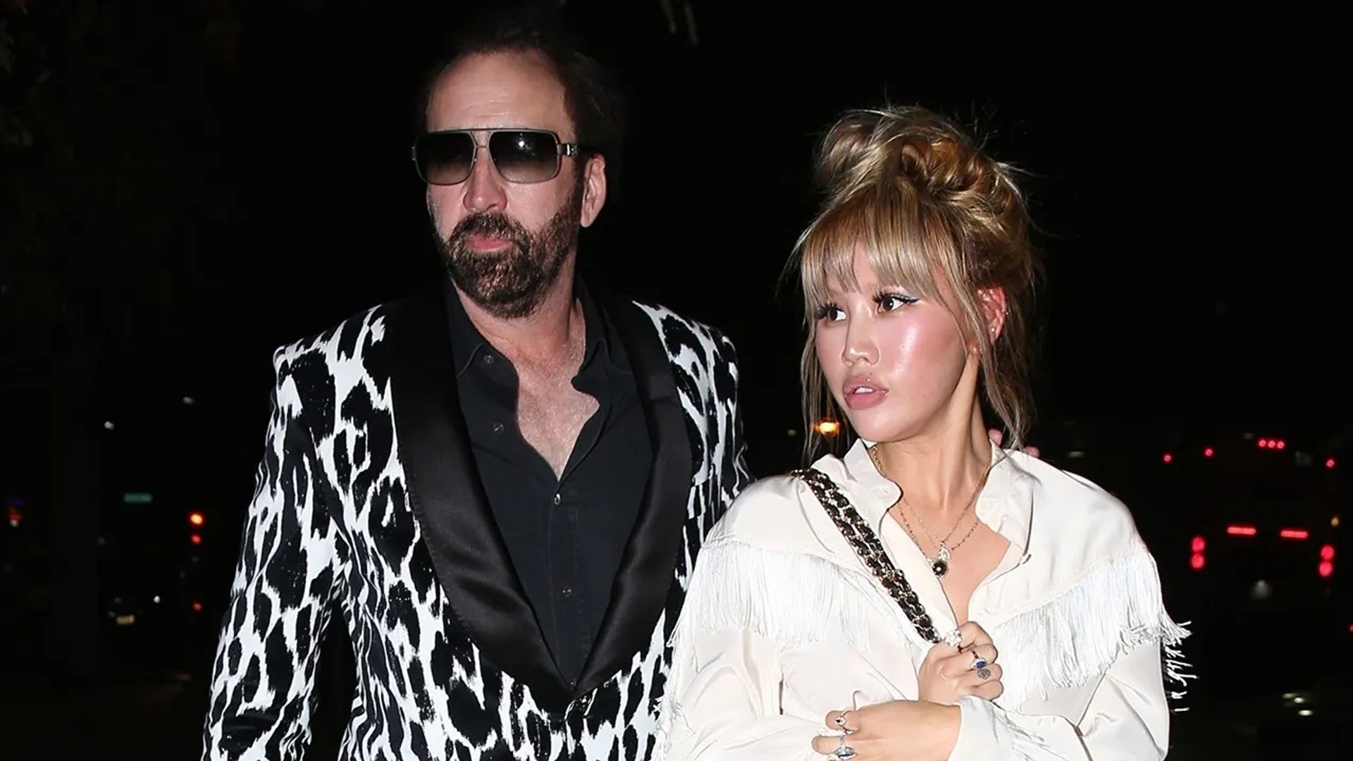 Nicolas Cage num blazer com padrão preto e branco e Erika Koike numa blusa branca a caminharem juntos à noite