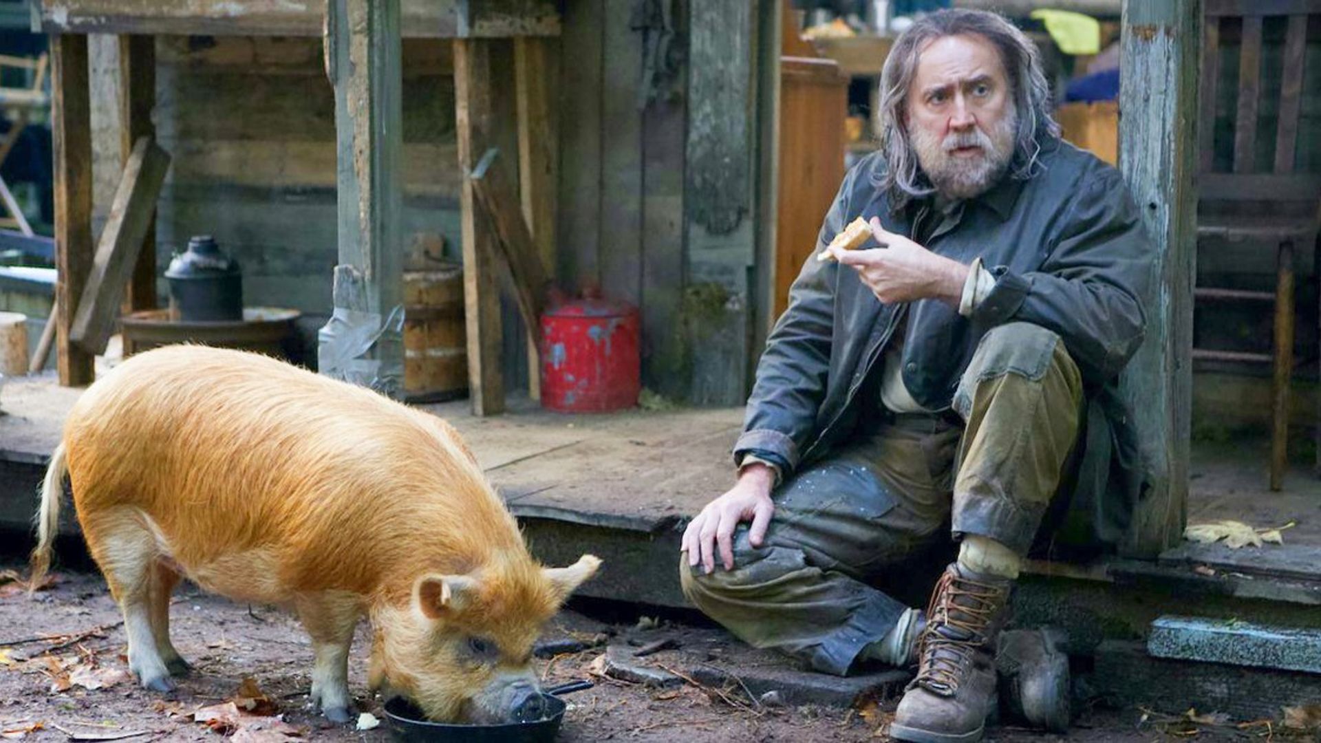Nicolas Cage com barba sentado na varanda de uma casa com comida na mão ao lado de um porco comendo numa tigela