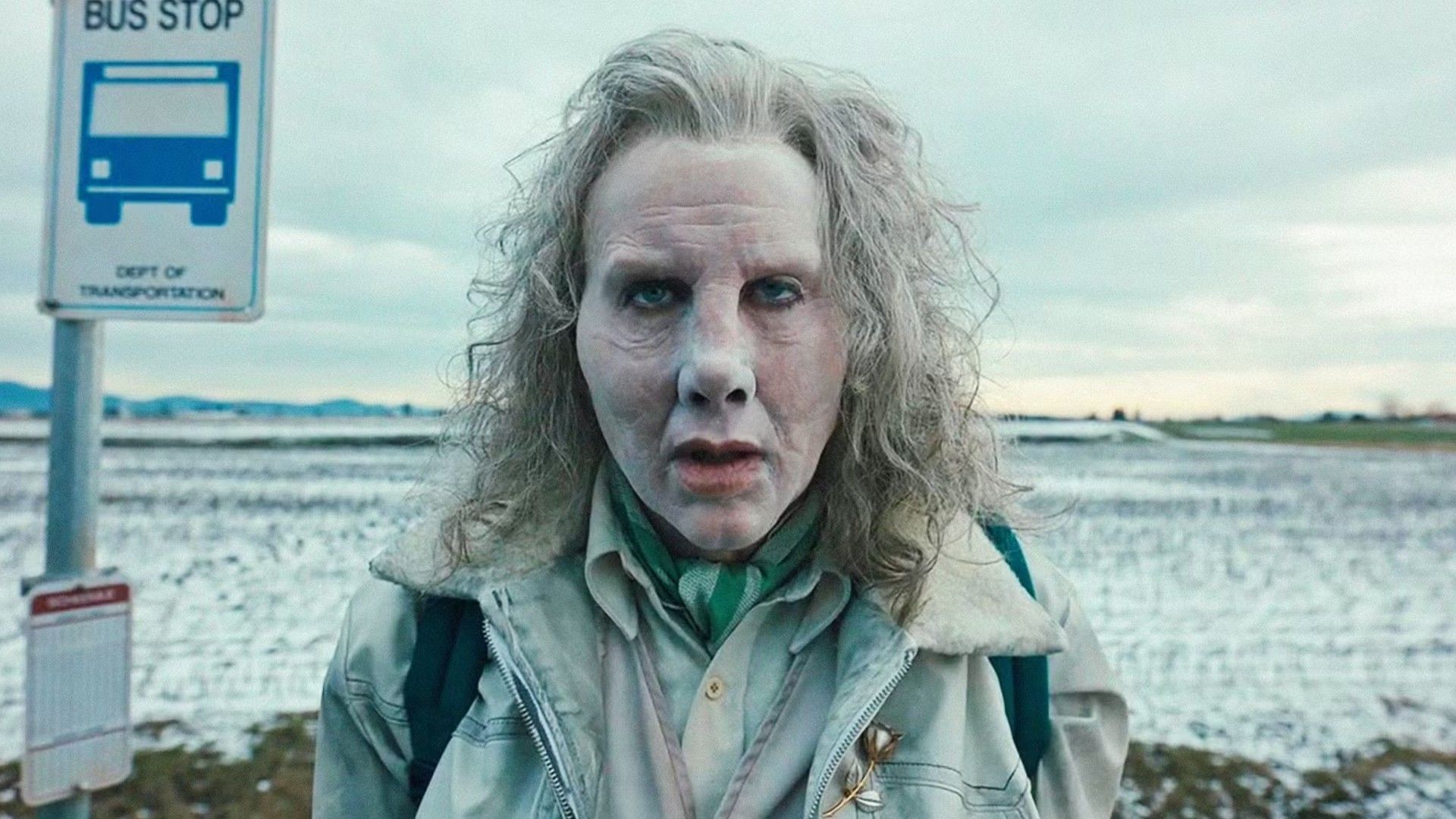 Nicolas Cage com cabelo grisalho e rosto pálido está junto a uma placa de paragem de autocarro contra um cenário de paisagem invernal