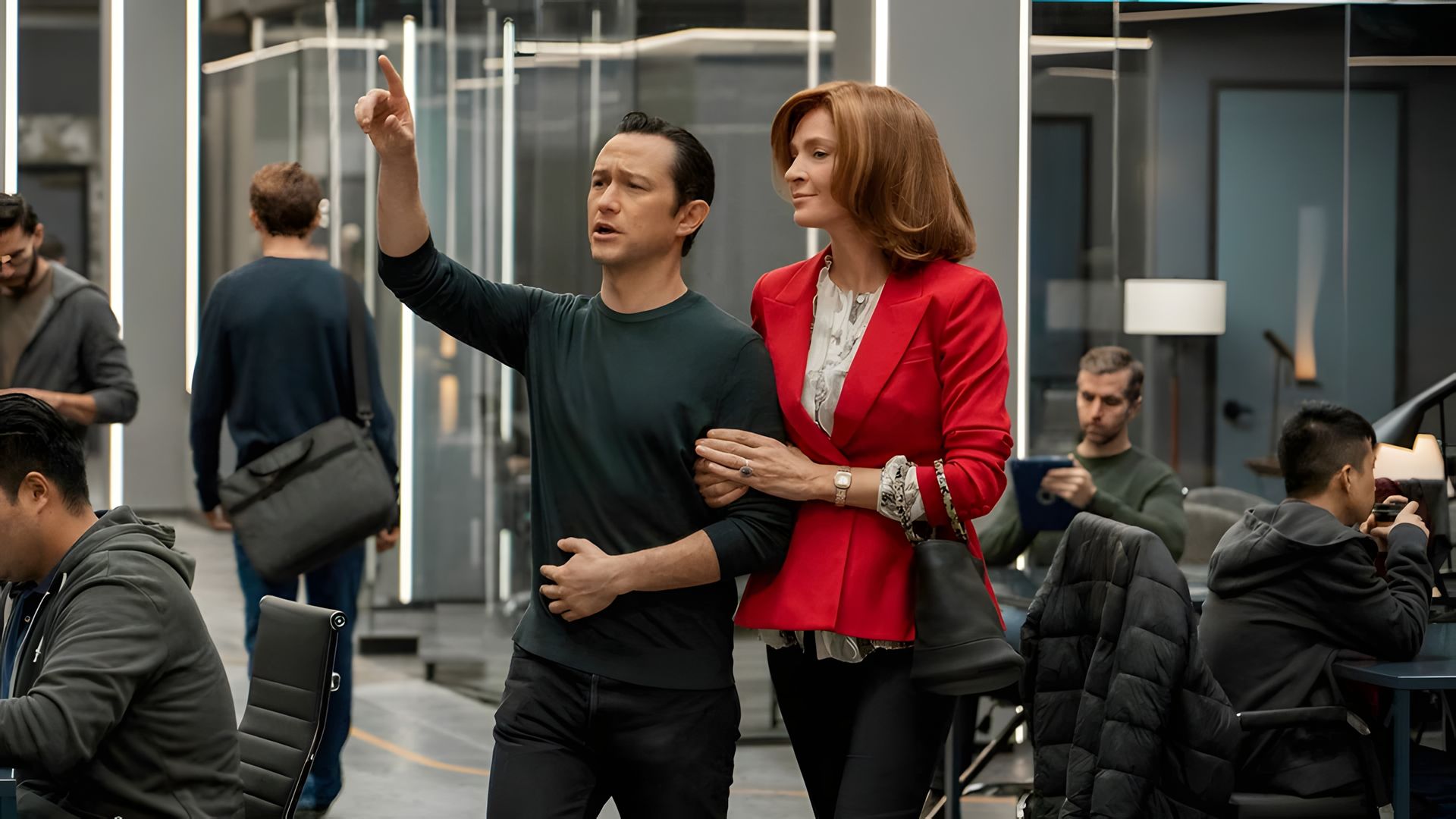 Joseph Gordon-Levitt vestido de escuro aponta para a frente enquanto Uma Thurman, de casaco vermelho, se agarra ao seu braço