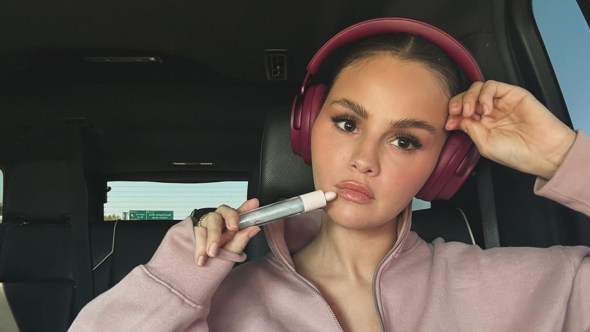 Selena Gomez numa camisola cor-de-rosa e auscultadores sentada num carro, segurando um produto cosmético junto ao rosto