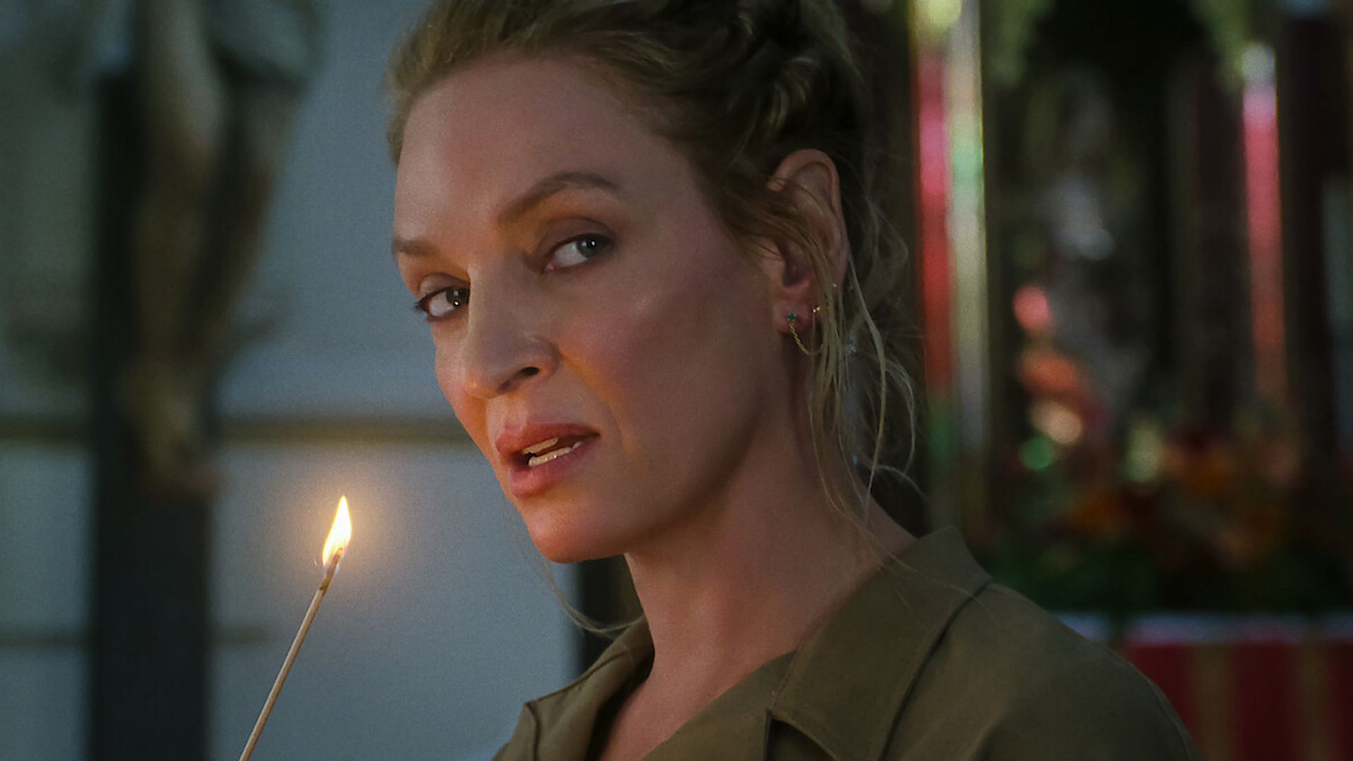 Uma Thurman com o cabelo apanhado segura uma vela acesa e olha para o lado com um fundo ligeiramente desfocado