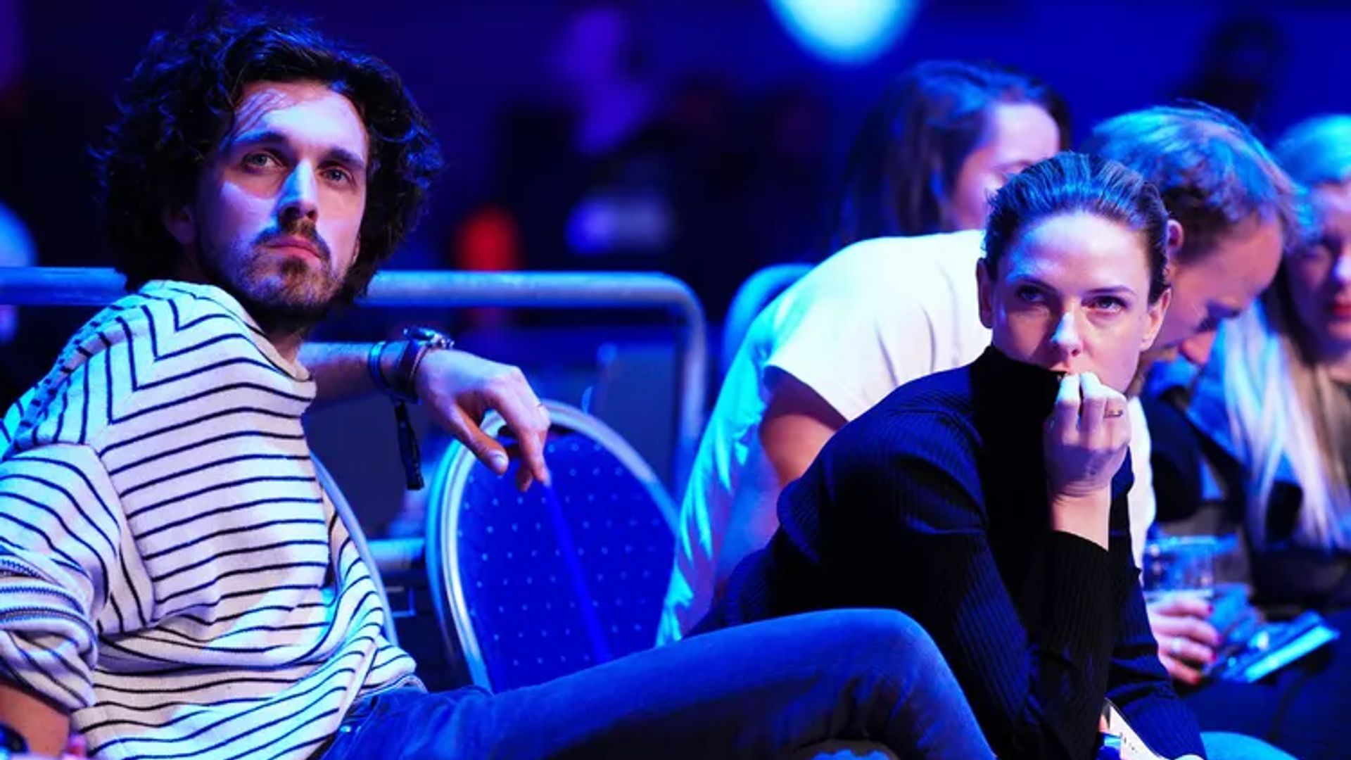 Rebecca Ferguson e Rory St. Clair Gainer sentados num hall com iluminação néon