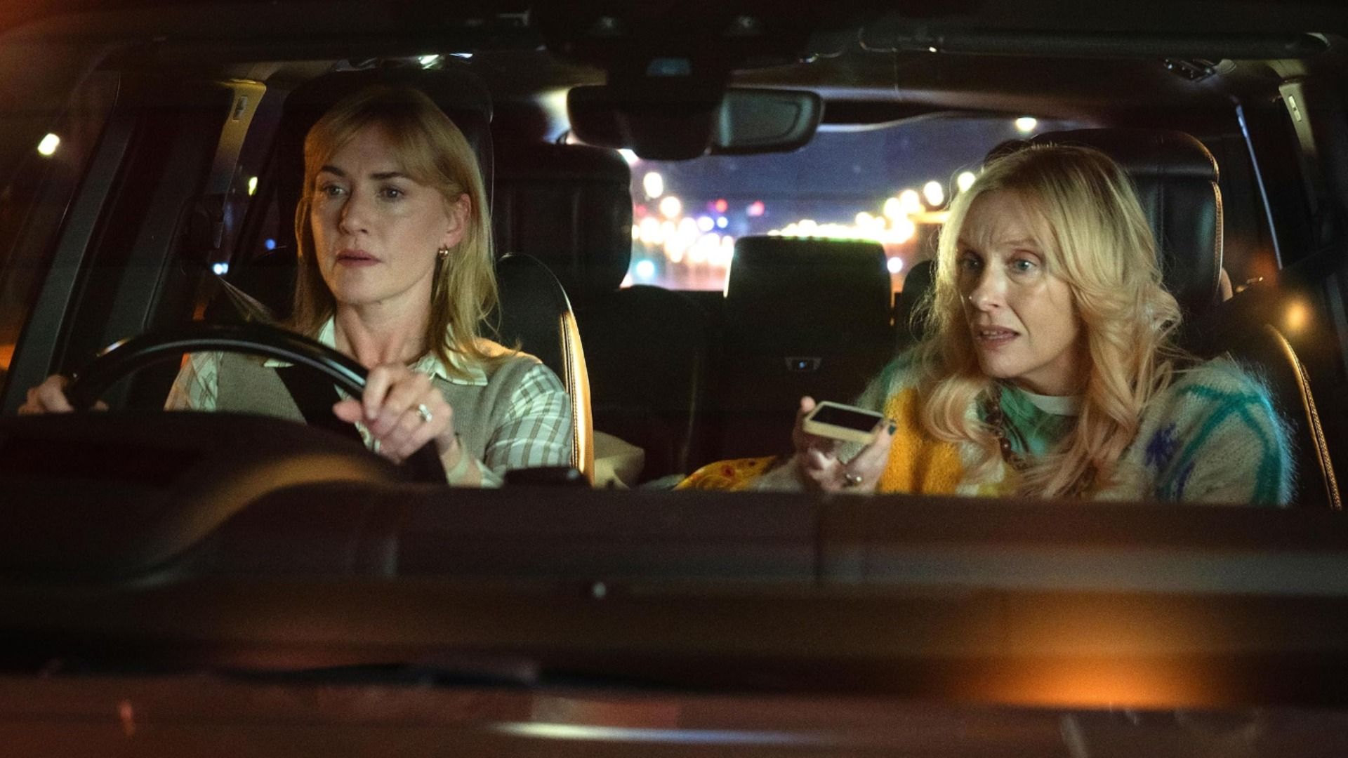 A atriz Kate Winslet senta-se ao volante de um carro enquanto a atriz Toni Collette fala ao telefone ao seu lado