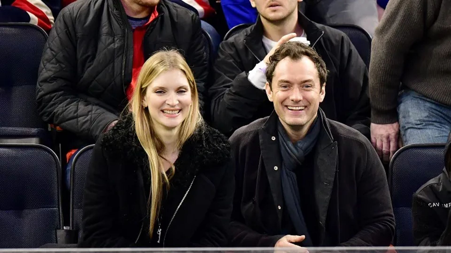 Jude Law e Phillipa Coan sentam-se lado a lado nas bancadas, sorrindo e olhando para a câmara