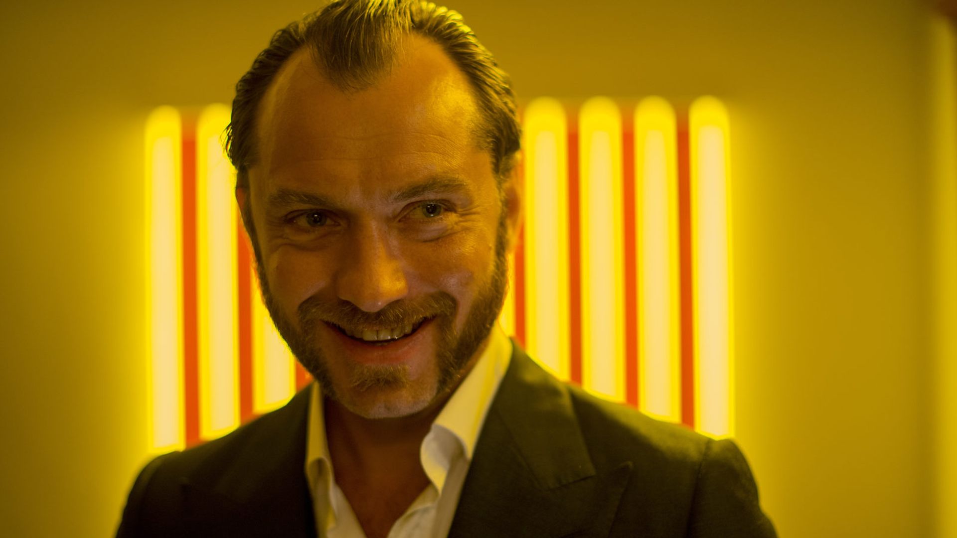 Jude Law de casaco sorri contra um fundo de listras de luz amarela brilhante