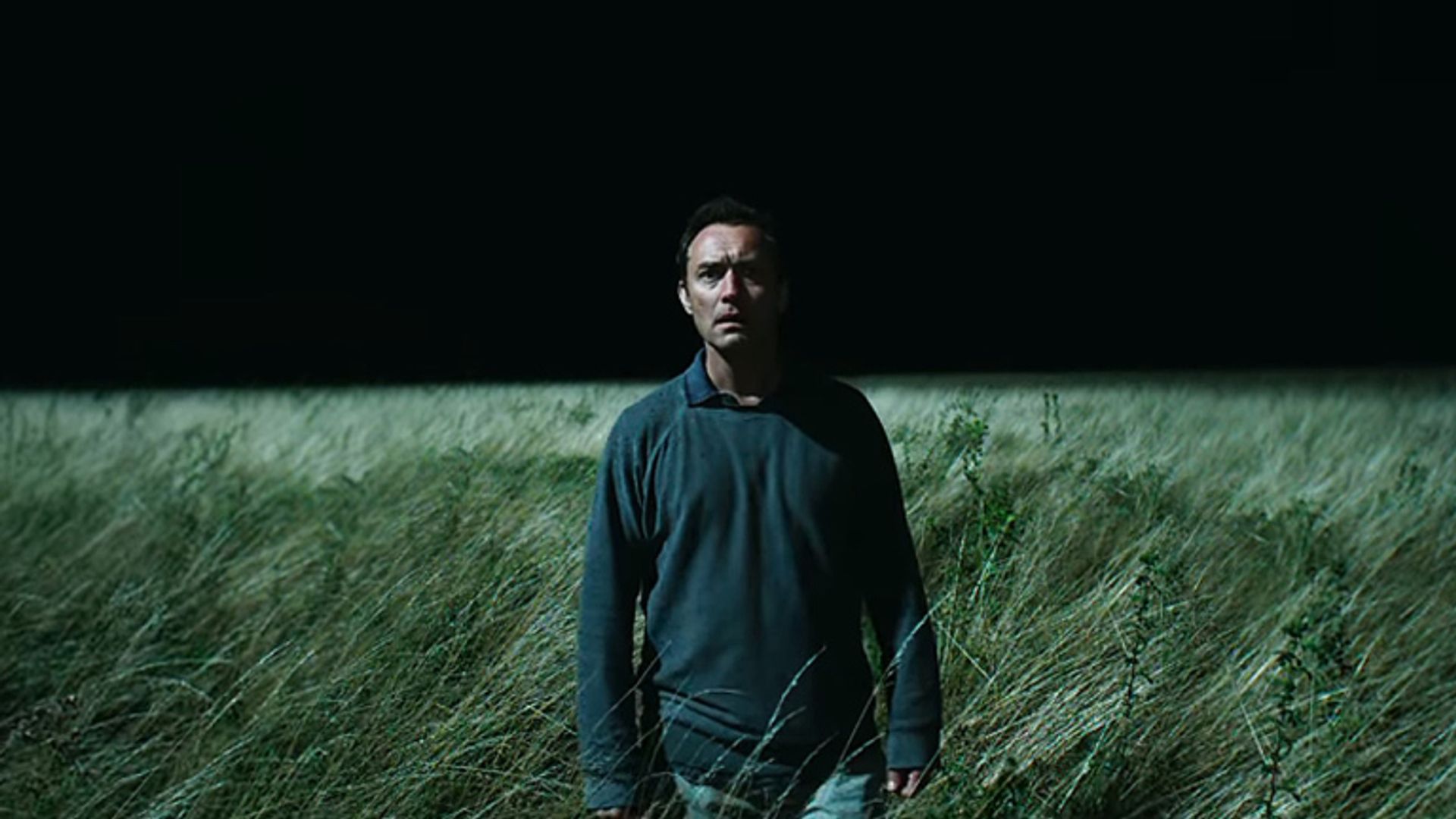 Jude Law com roupa escura está num campo de erva alta à noite, iluminado por luz intensa