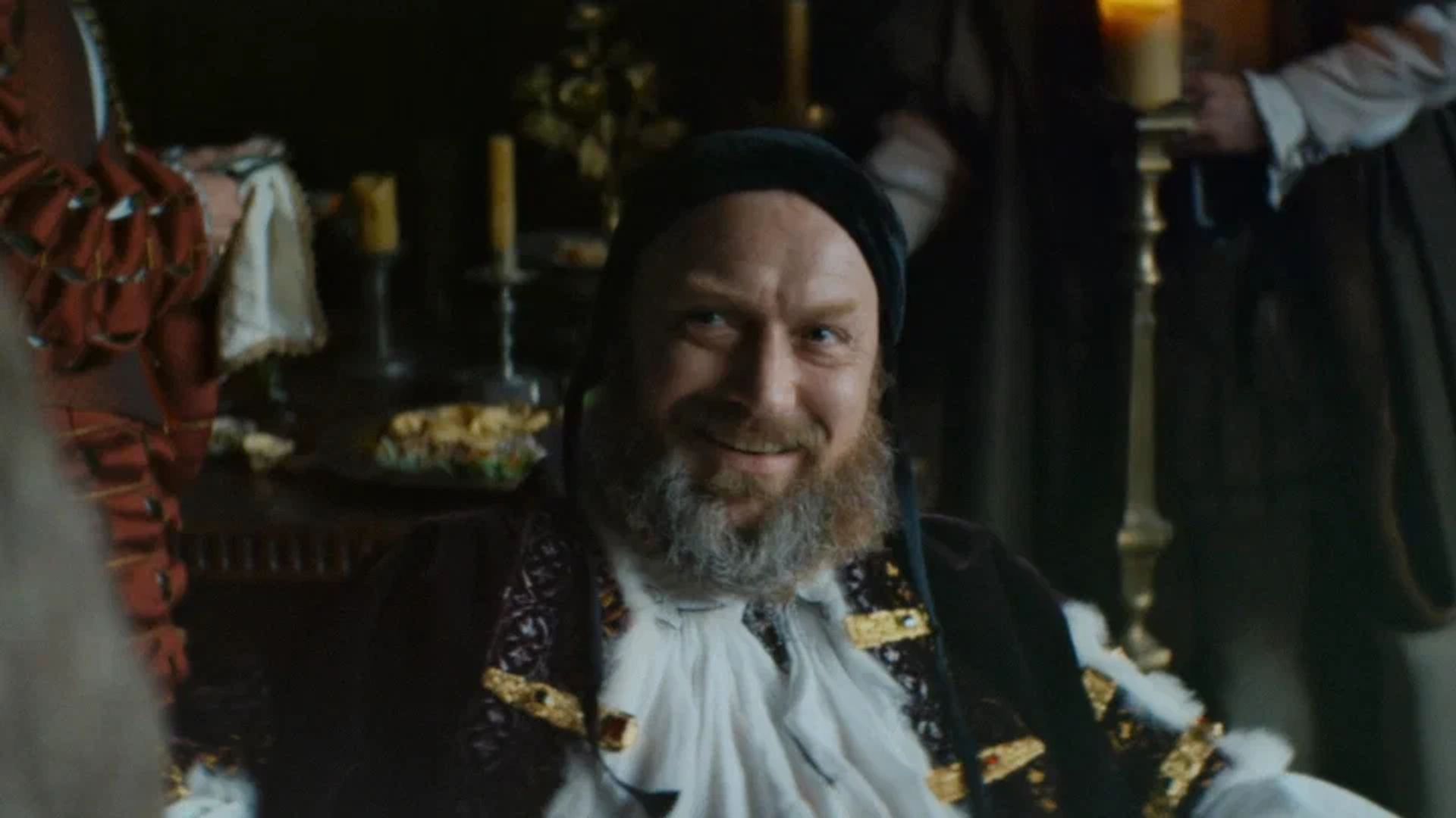 Jude Law em traje medieval com barba sorri enquanto está sentado numa mesa festiva iluminada por velas
