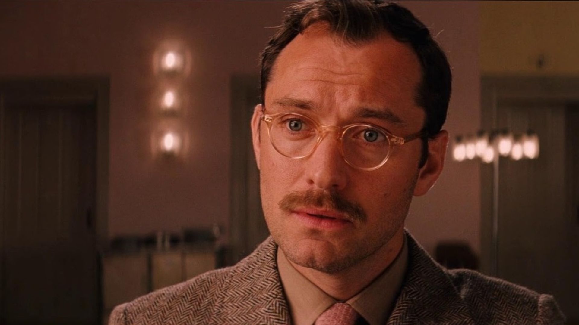 Jude Law usando óculos e bigode, de fato, olha com um olhar sério no interior acolhedor de uma sala