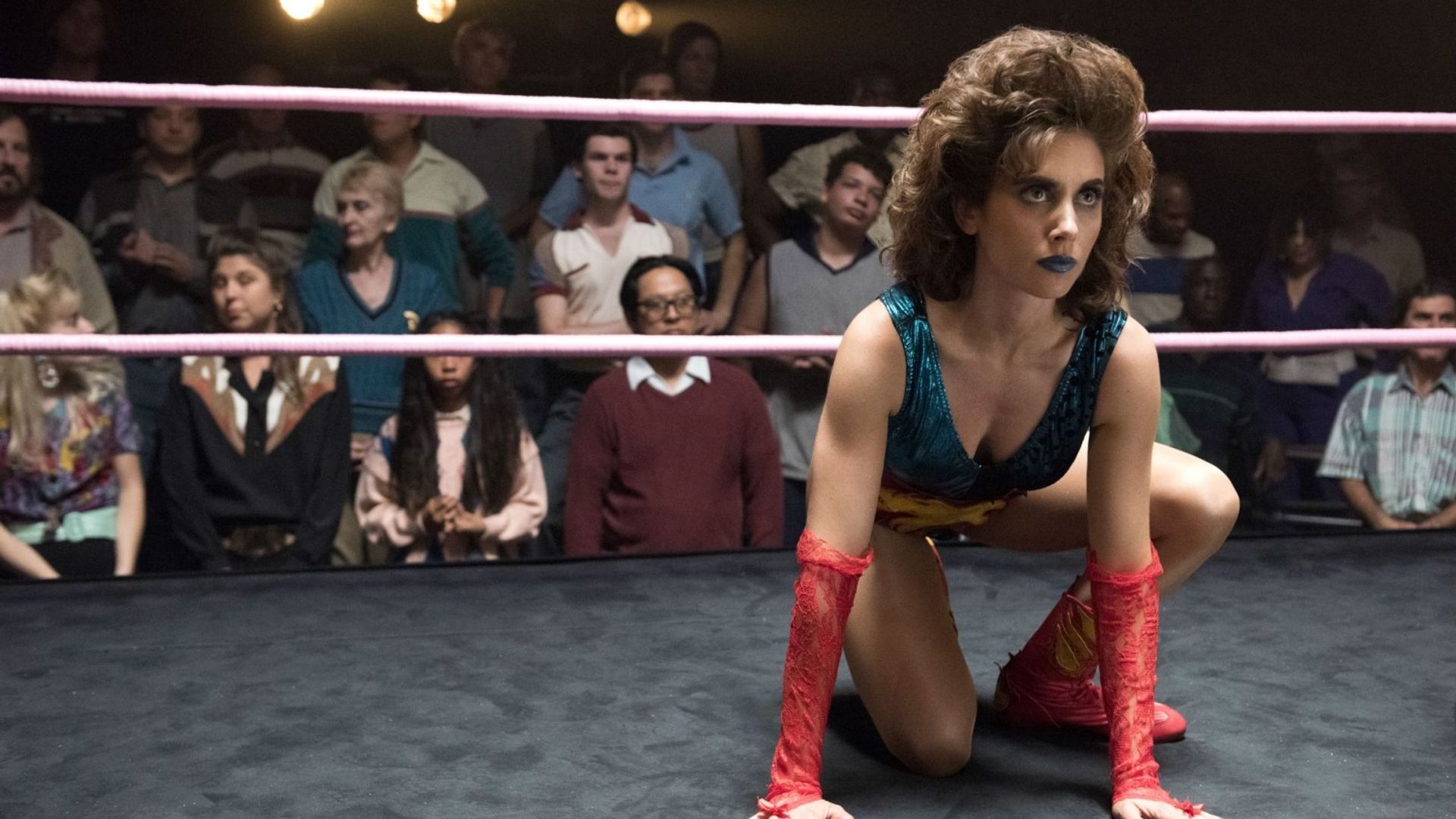 Alison Brie em fato de lutadora senta-se numa posição no ringue
