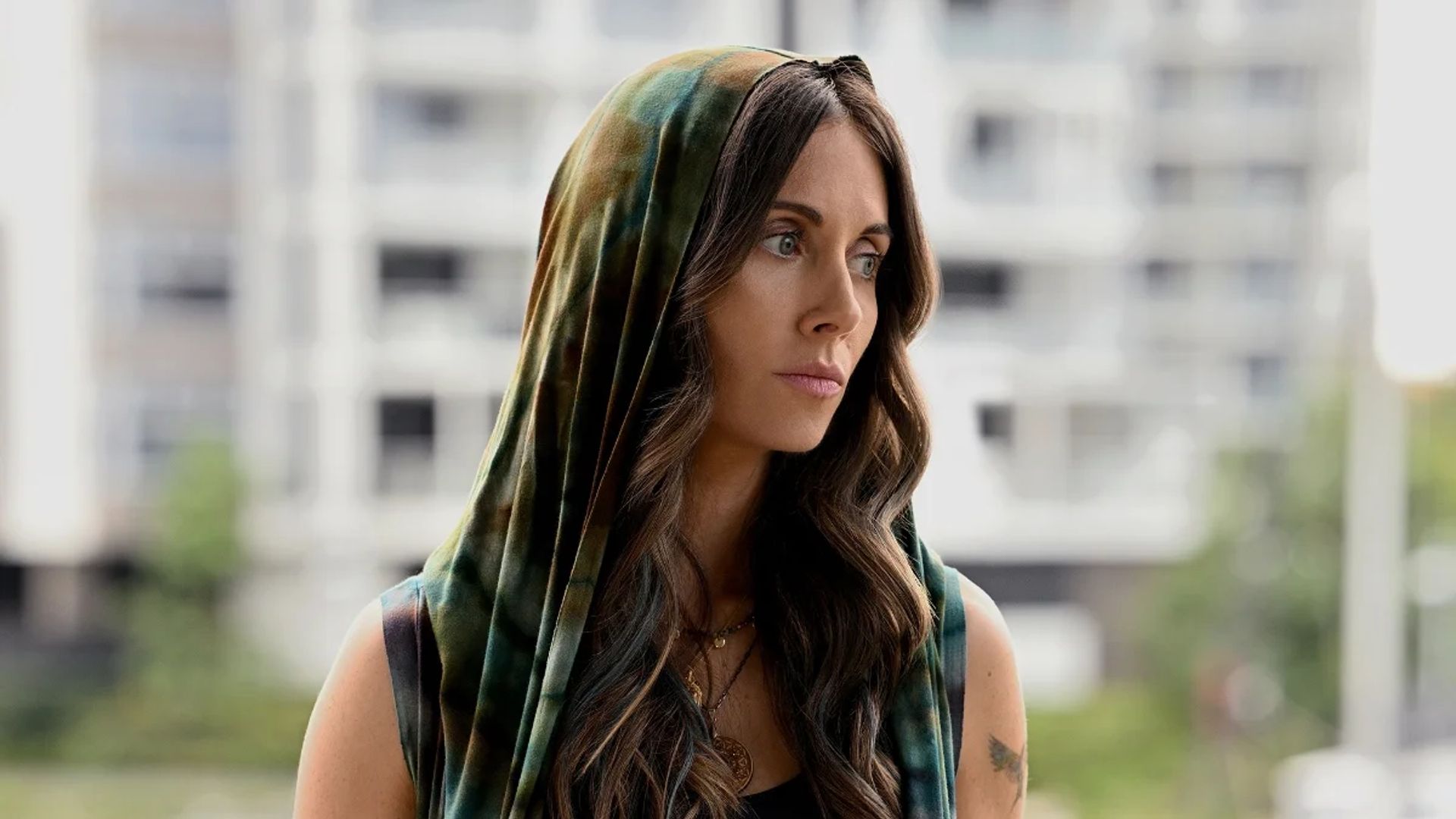 Alison Brie com cabelo comprido e capuz olha pensativamente para o lado contra o cenário de um edifício alto