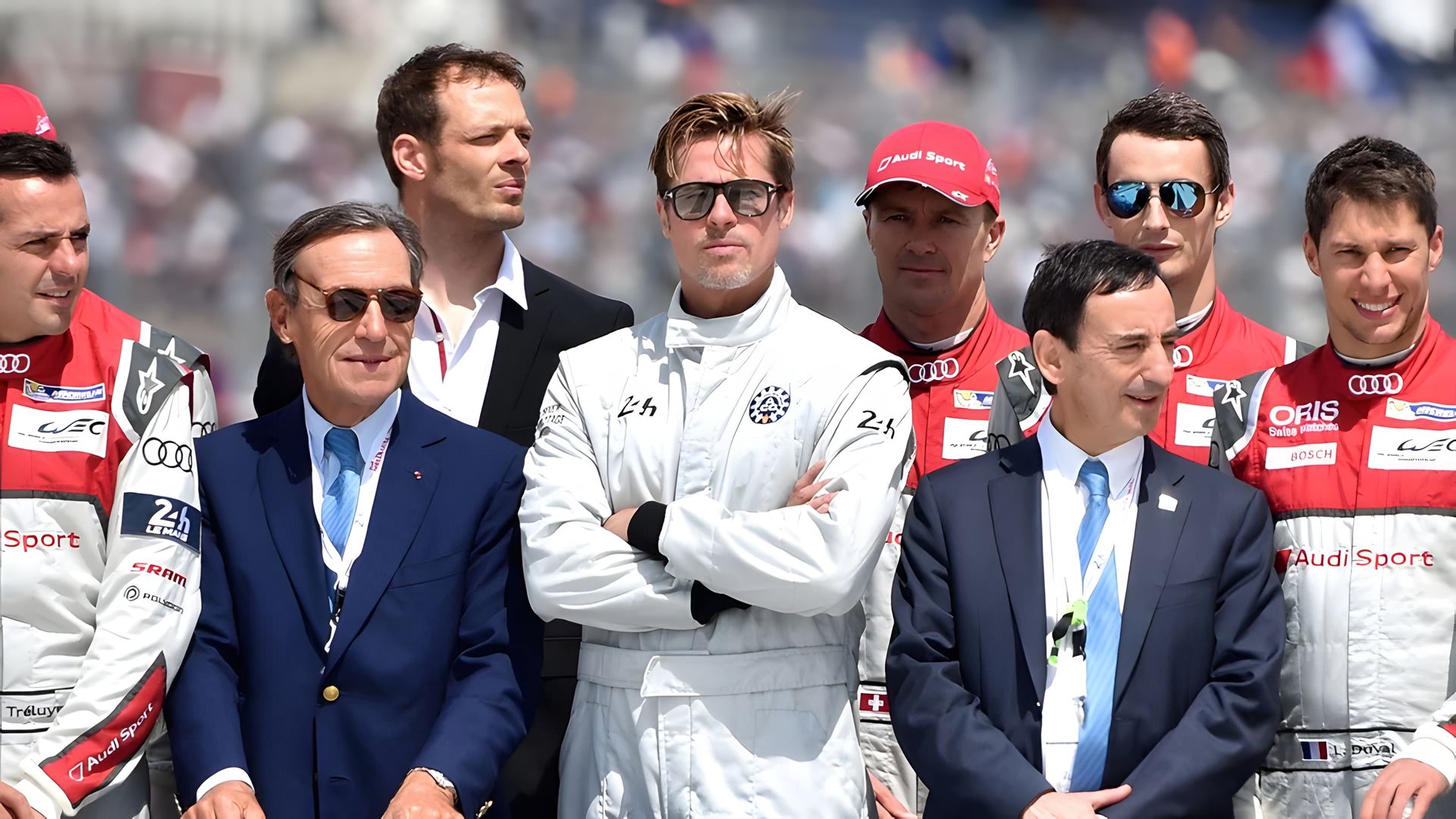 Brad Pitt em equipamento de piloto de corridas está de pé rodeado pela sua equipa