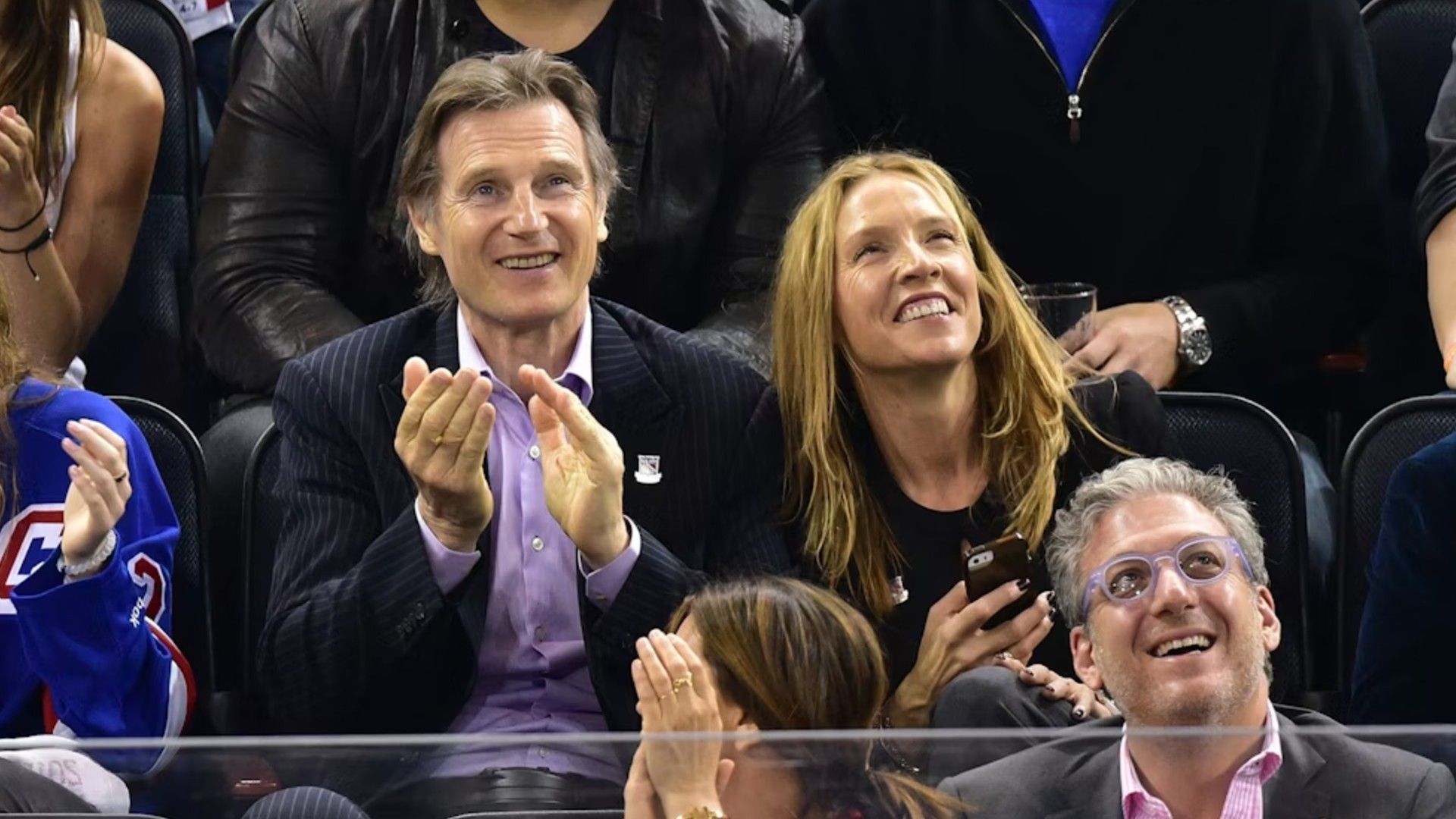 Liam Neeson e Freya St. Johnston sentados nas bancadas durante um jogo desportivo e a torcer com alegria