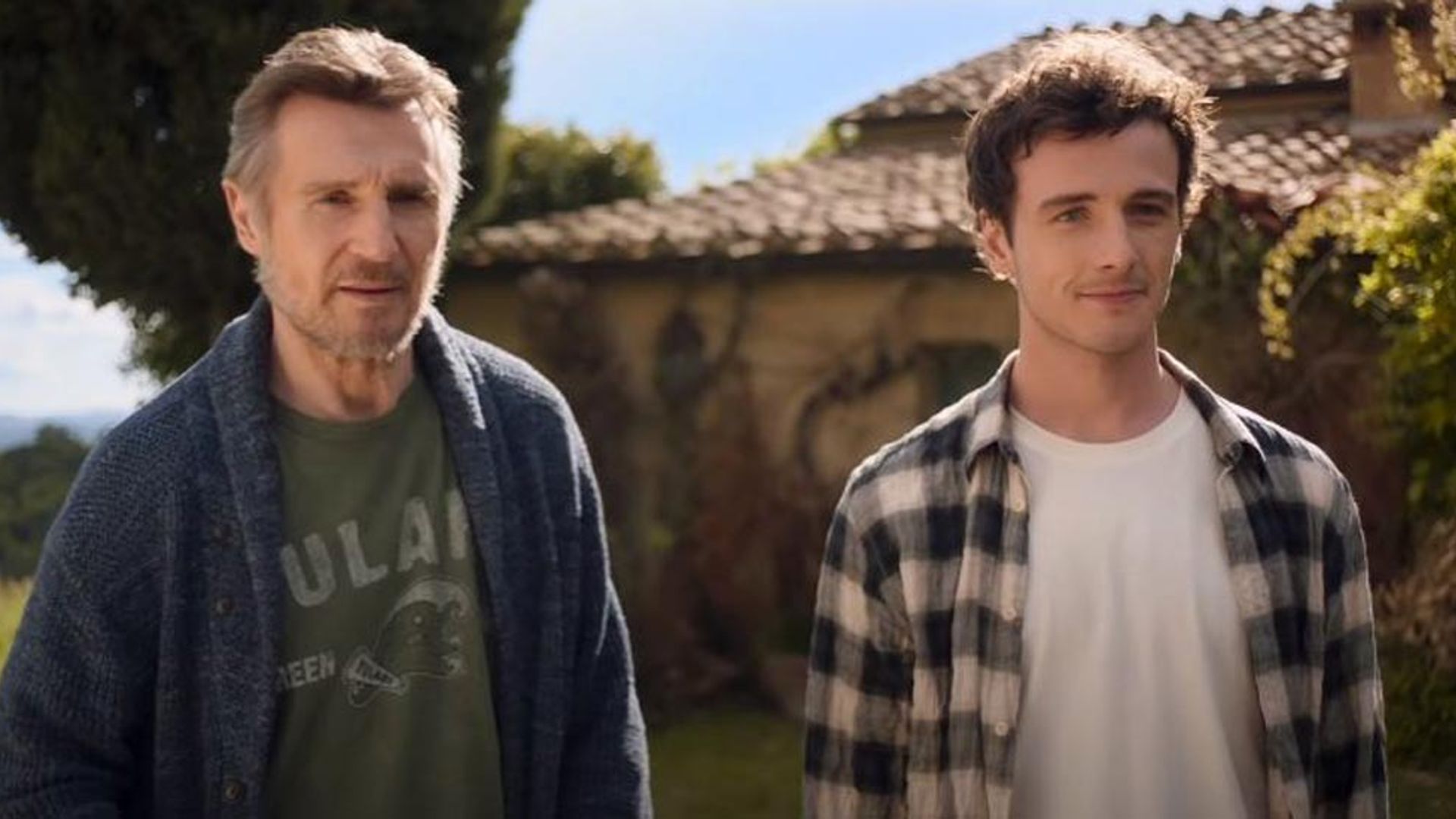 Liam Neeson e Michael Richardson estão lado a lado em frente a uma casa com telhado de telhas num dia ensolarado