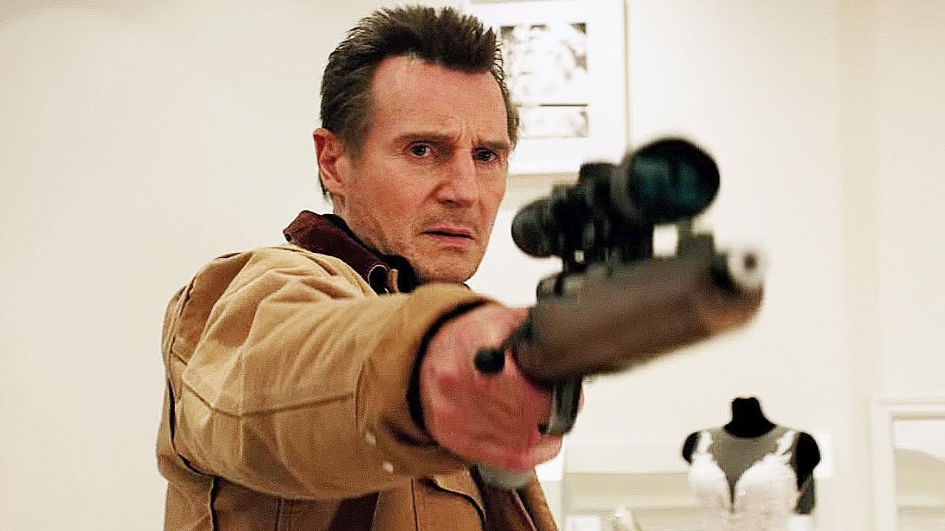 Liam Neeson numa jaqueta bege aponta uma espingarda com mira telescópica, com um manequim de vestido de noiva atrás dele