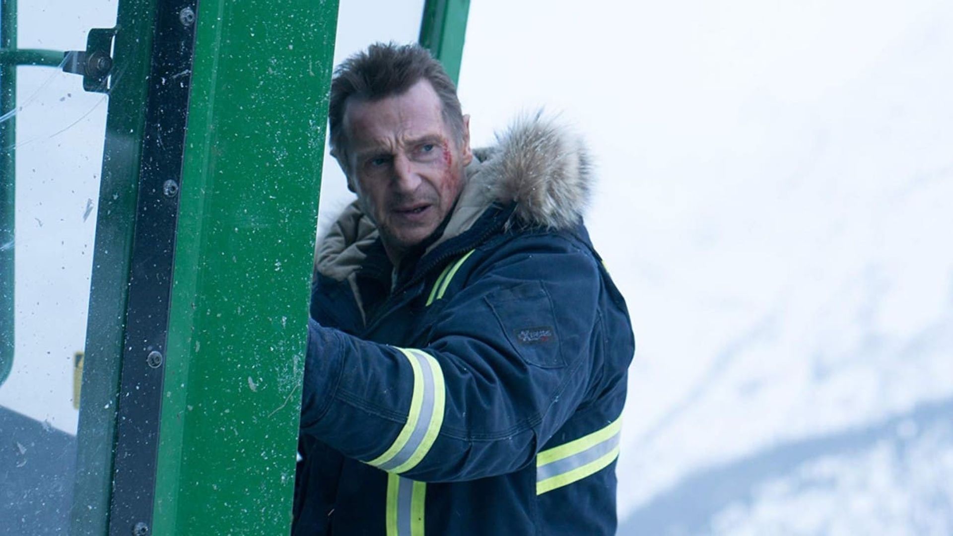 Liam Neeson numa jaqueta de inverno com capuz agarra a porta de uma cabana verde com uma paisagem coberta de neve ao fundo