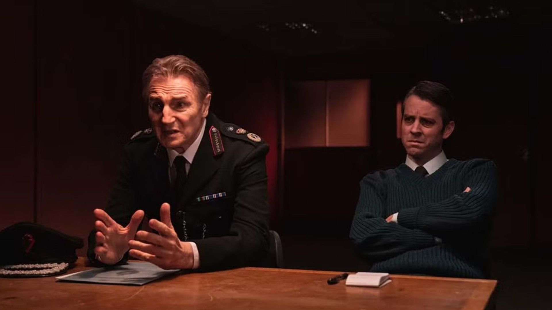 Liam Neeson em uniforme policial sentado numa mesa de interrogatório, cena da série "Derry Girls"
