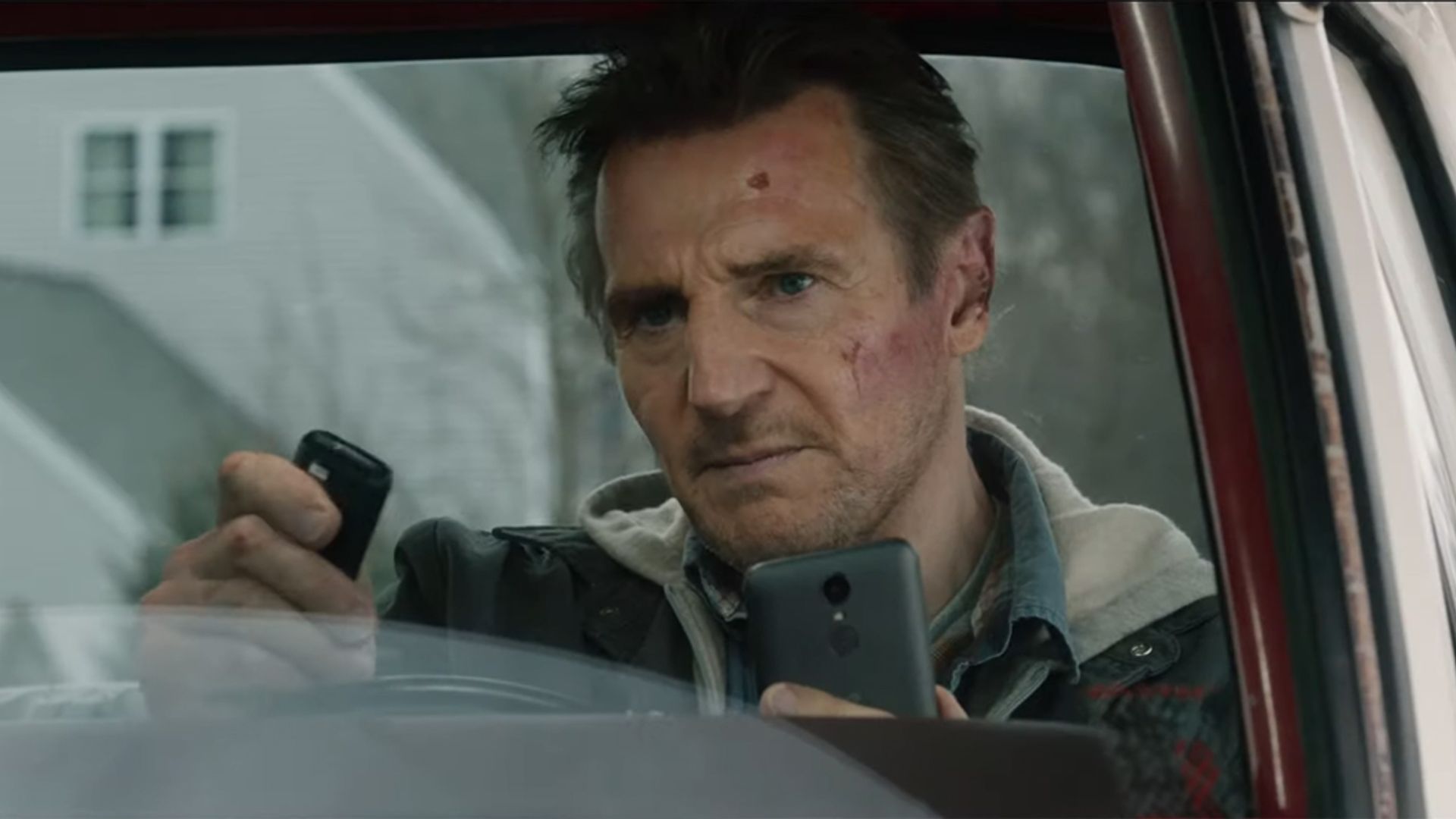 Liam Neeson está sentado num carro olhando tensamente para o telefone e controlo remoto nas suas mãos