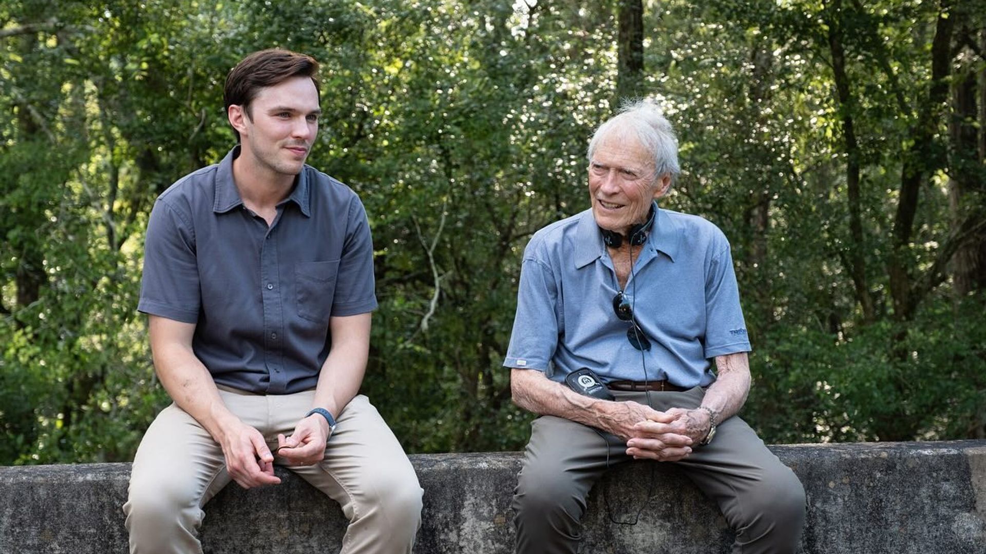 Nicholas Hoult e Clint Eastwood em roupa casual sentados numa barreira de betão a conversar com uma floresta ao fundo