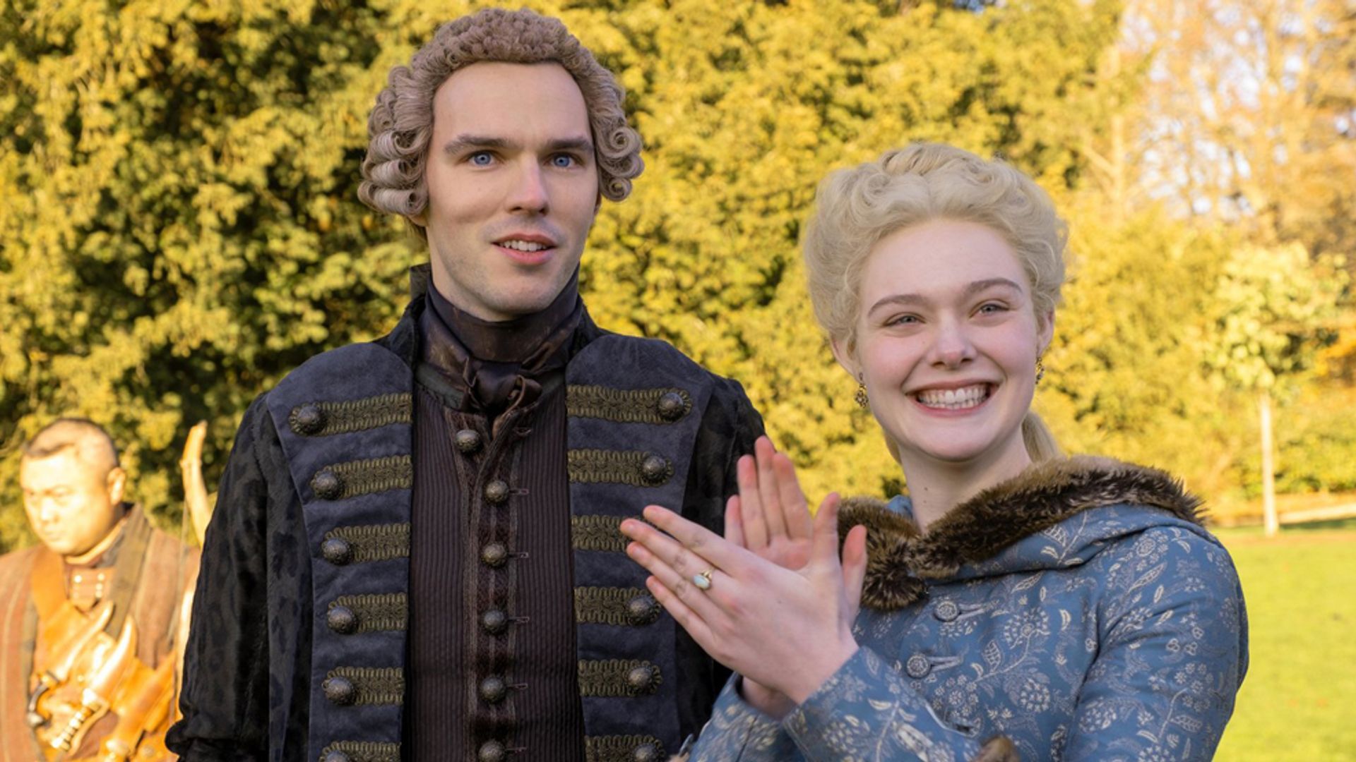 Nicholas Hoult e Elle Fanning com trajes de época do século XVIII, de pé na rua, a atriz está a sorrir e a bater palmas